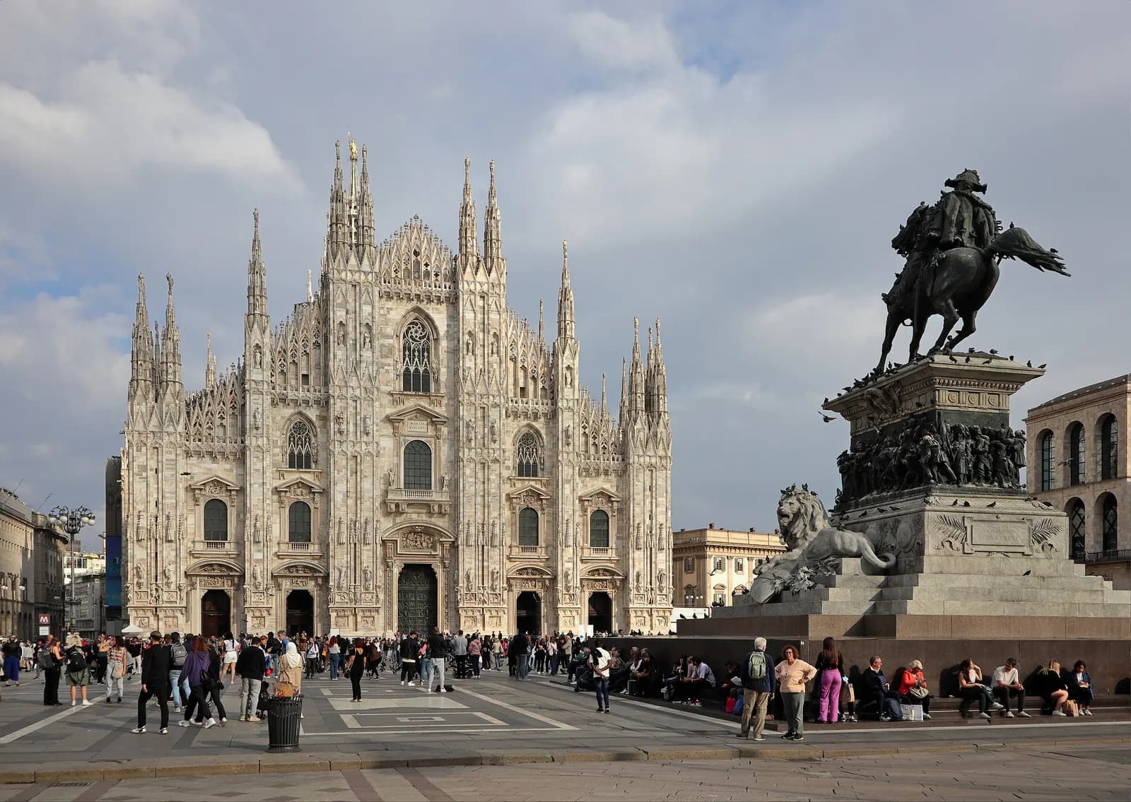 The white marble facade of Milan's Duomo in afternoon sun, with the equestrian monument to Vittorio Emanuele II and people crossing Piazza del Duomo.