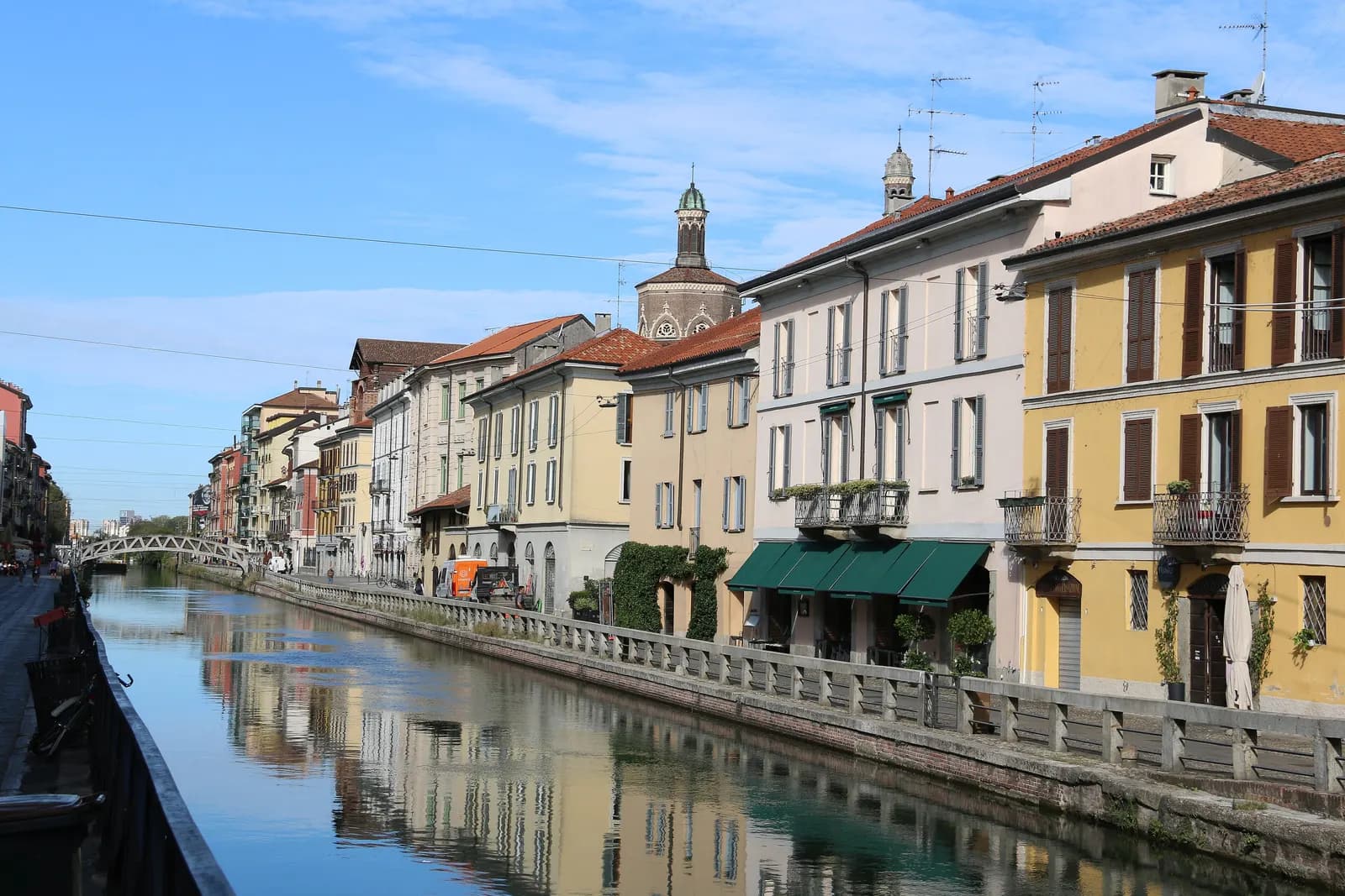Pastel houses with green awnings line the Naviglio Grande, reflected in the still water of the canal, with an iron footbridge further along.