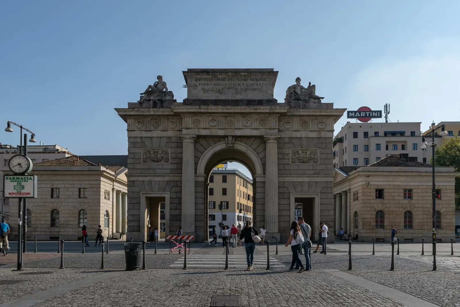 The neoclassical Porta Garibaldi arch and its two toll houses in Piazza XXV Aprile, at the south end of Corso Como, with people crossing the cobbled square.