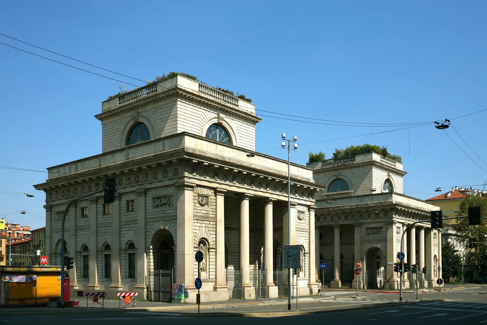 The two neoclassical toll houses of Porta Venezia at a road junction under a clear blue sky, with traffic lights and a metro sign.