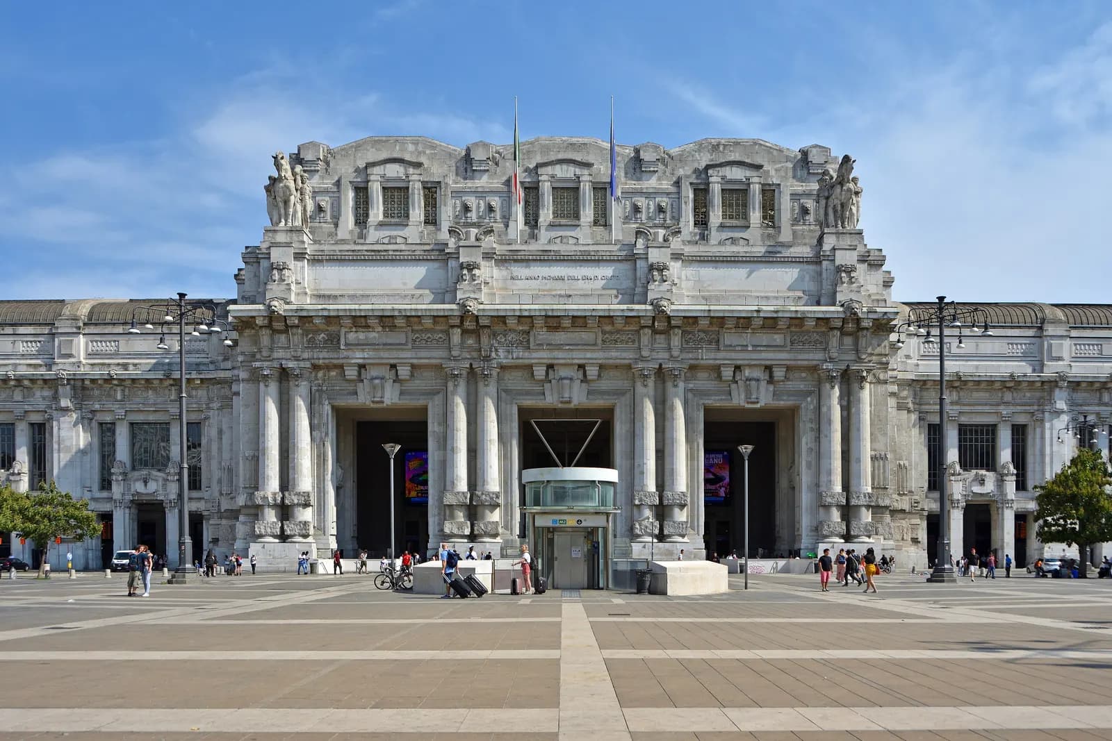The monumental stone facade of Milano Centrale station behind the wide Piazza Duca d'Aosta, with travellers pulling suitcases towards the entrance.