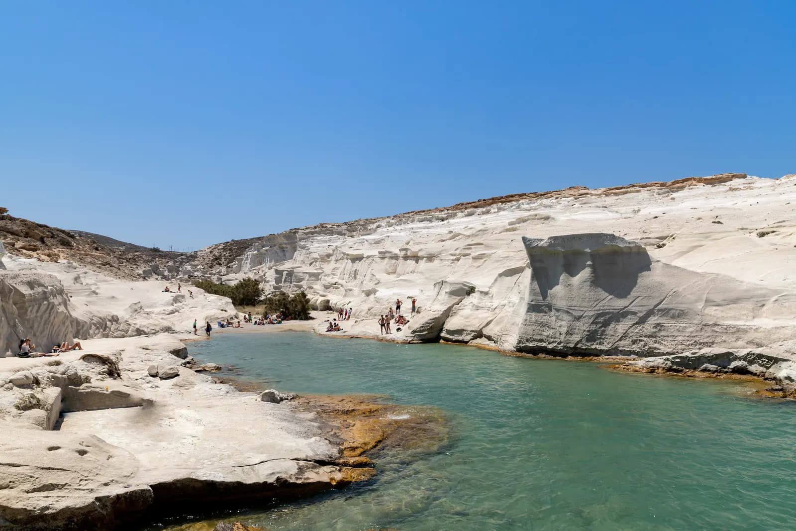 The white volcanic rocks of Sarakiniko around a narrow inlet of green water, with bathers on the rocks