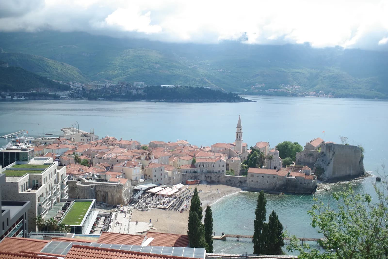 Budva old town on its small peninsula, with the bell tower, the citadel walls and a sand beach, and hills across the water behind