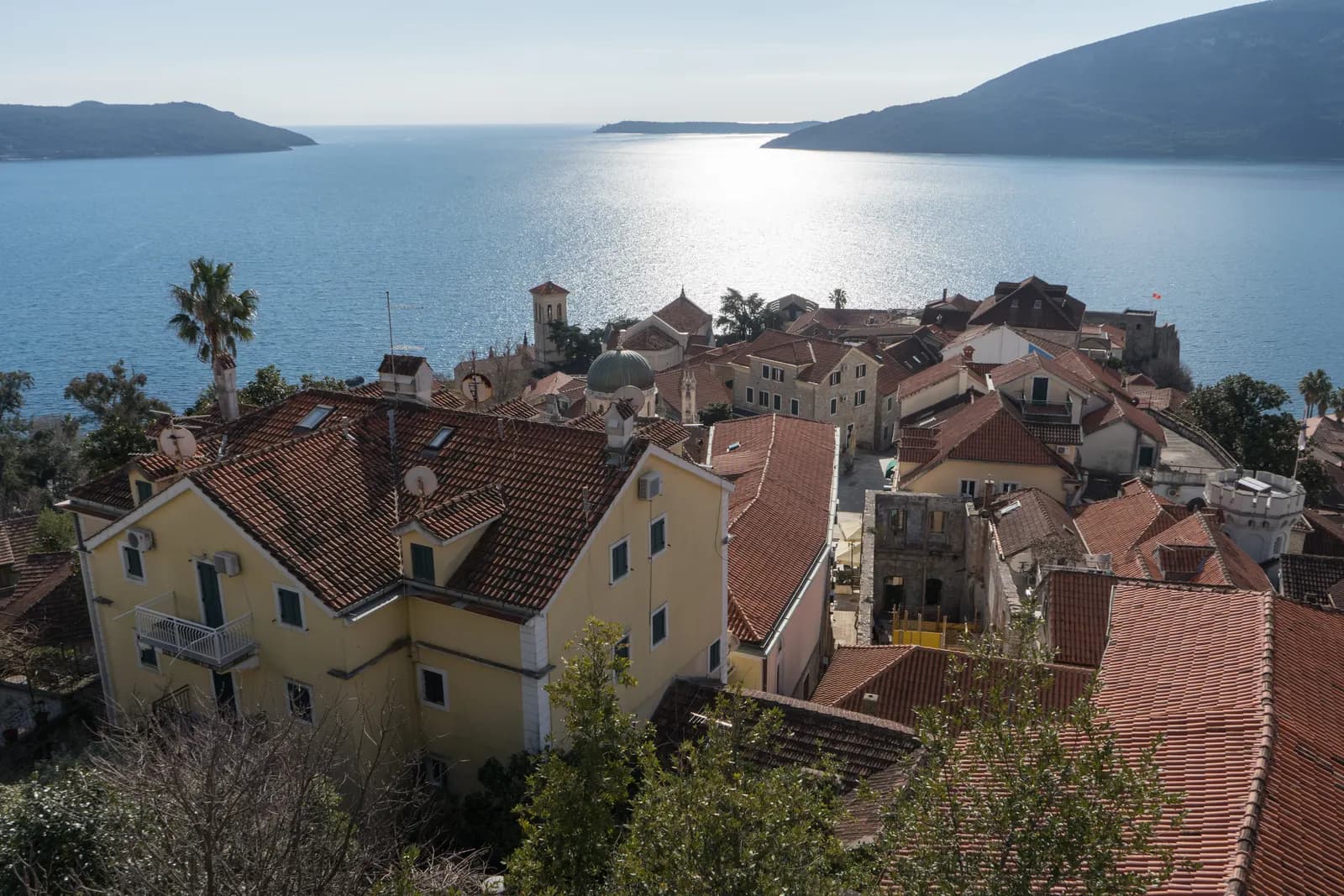 Tiled roofs of Herceg Novi old town seen from the Kanli Kula fortress, with a stone tower and a domed church, and the sea and headlands beyond
