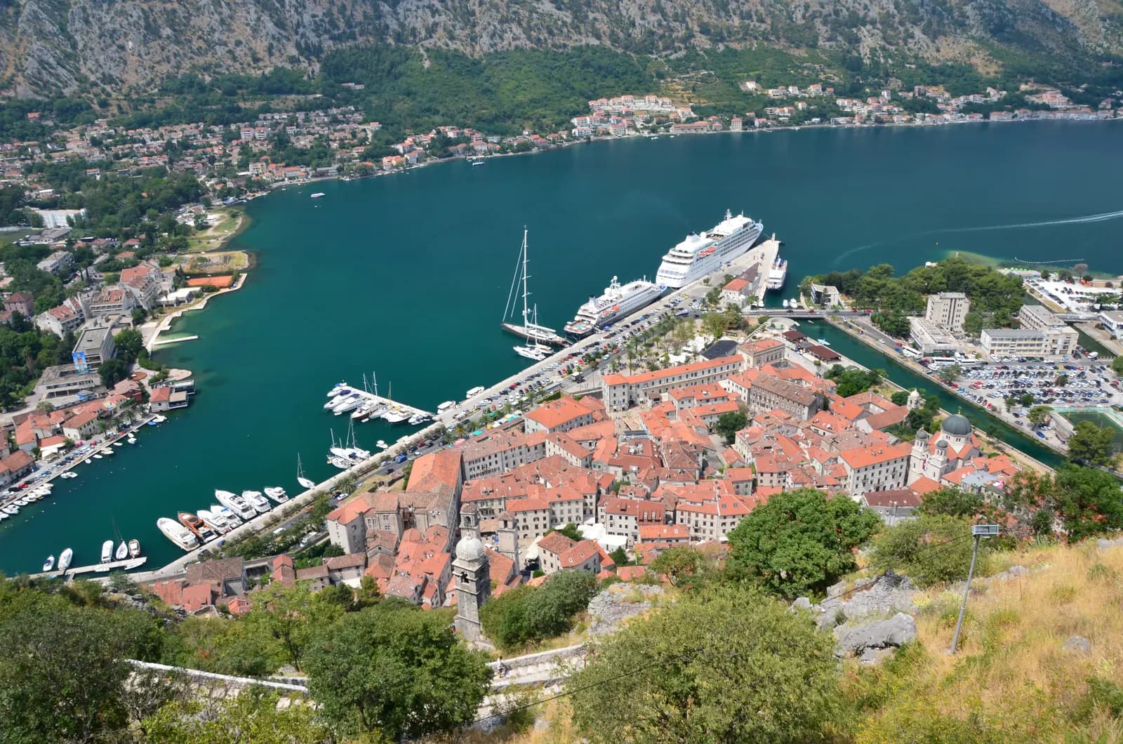 Kotor old town seen from the fortress path: red roofs inside the walls, cruise ships and cars along the quay, and the bay with villages below the mountains