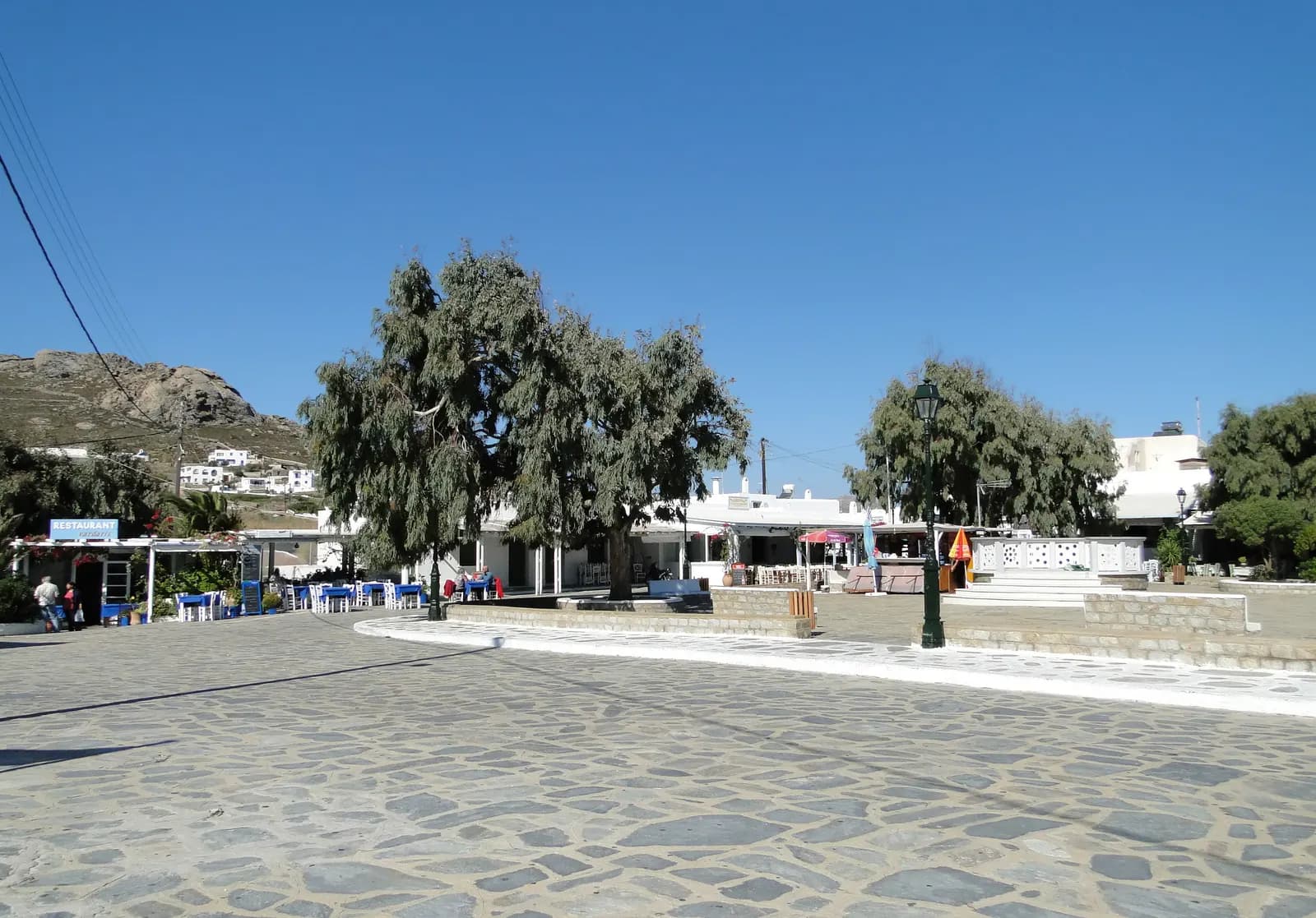 The paved central square of Ano Mera with large trees, taverna tables and low white buildings under a clear sky