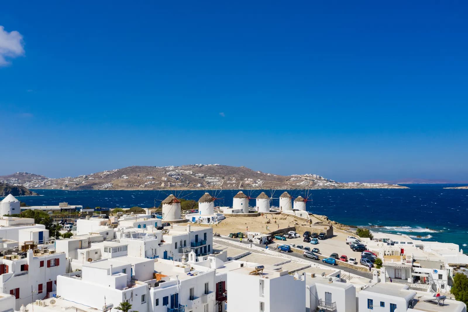 The row of white windmills on the hill above the rooftops of Chora, with cars parked along a road below them and the bay behind