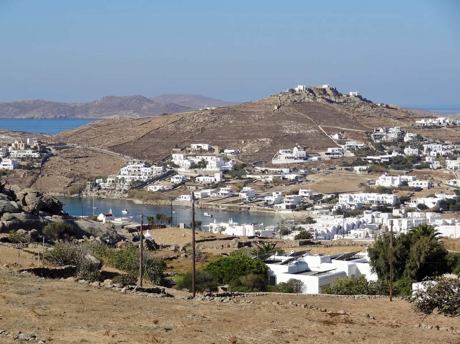 The bay of Ornos seen from the dry hillside above, with white houses and hotels around the beach and small boats in the water
