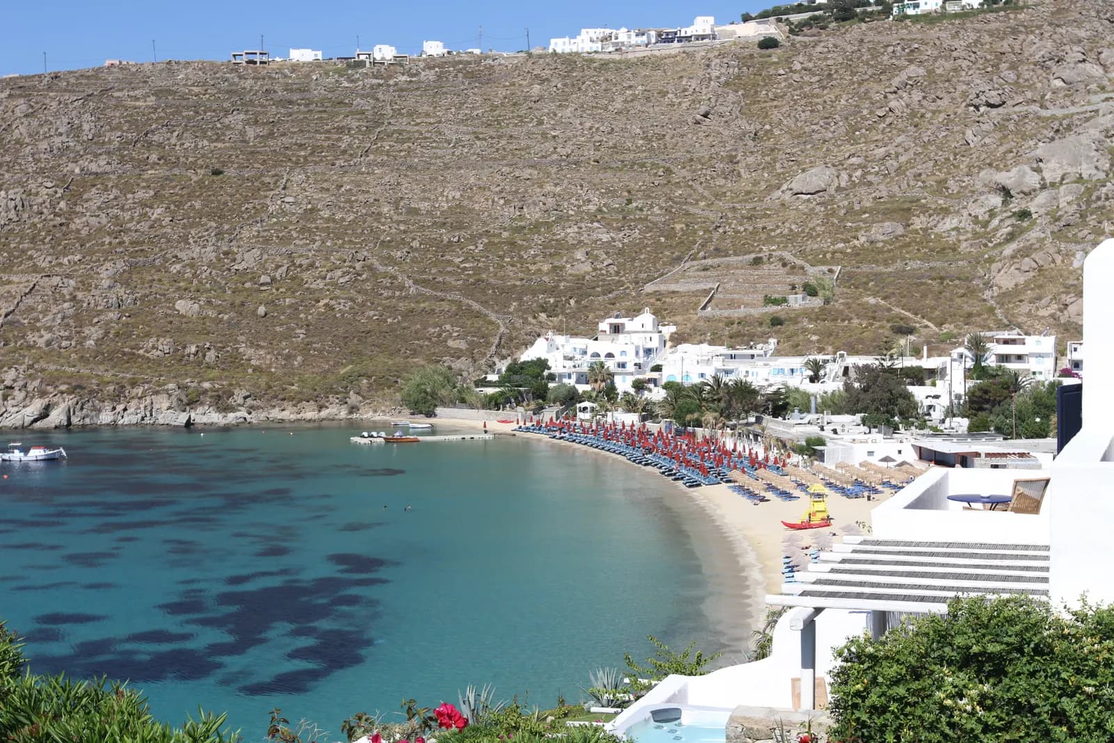 Psarou beach seen from a hotel terrace, a curve of sand lined with sunbeds and umbrellas below white buildings and a bare hillside