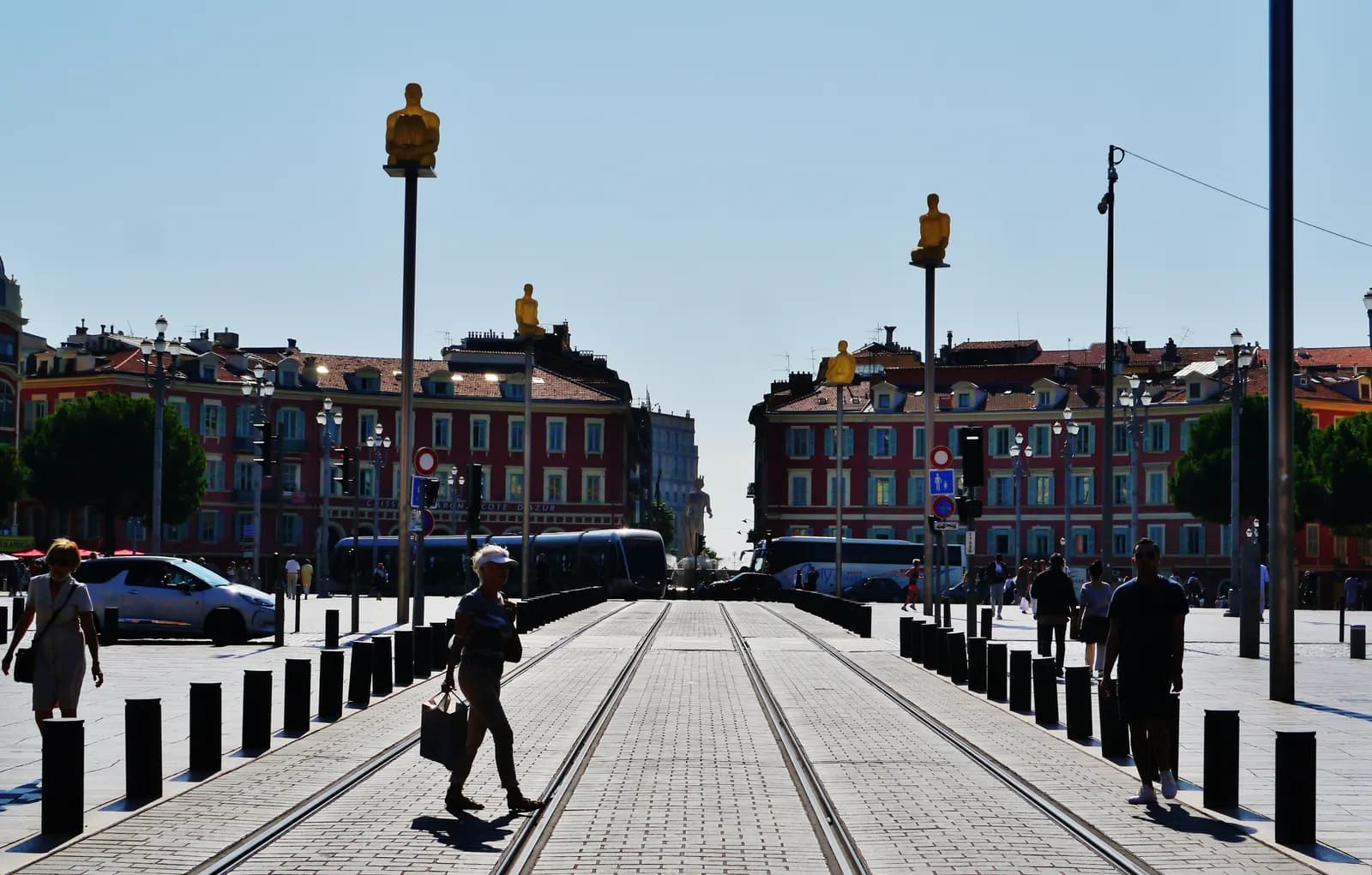 Tram tracks crossing the paved Place Masséna in Nice, with the red arcaded buildings of the square and the seated figures on their masts