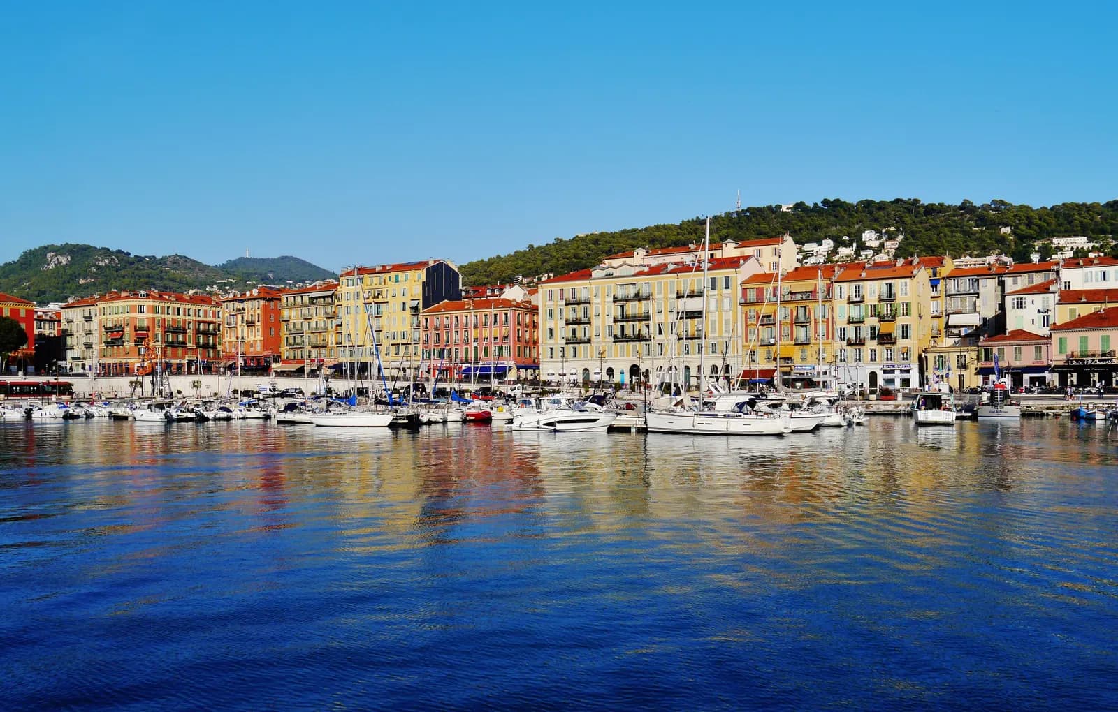 Yachts and fishing boats moored in Port Lympia in Nice, with the coloured house fronts of the quay and the wooded hill of Mont Boron behind