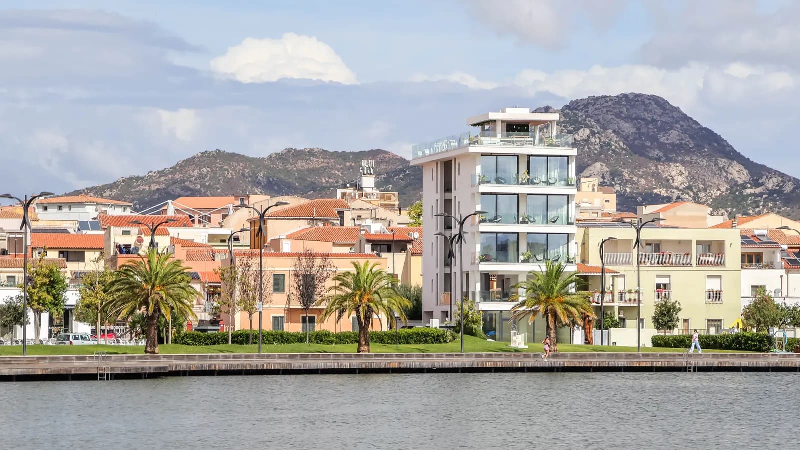 The Olbia waterfront seen across the inlet, with palms and street lamps along the promenade and the granite hills of Gallura behind the town