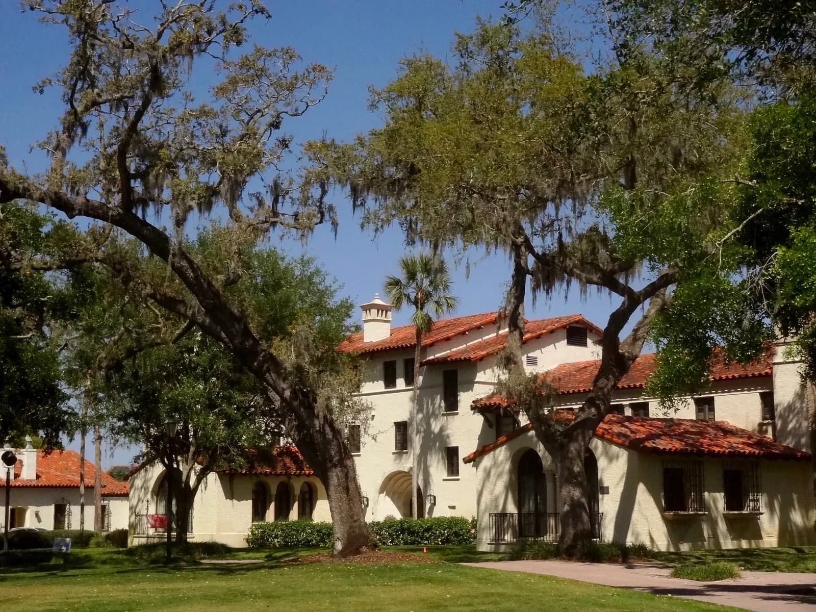 A cream Spanish-style building with red tile roofs under large oak trees hung with Spanish moss on the Rollins College campus in Winter Park