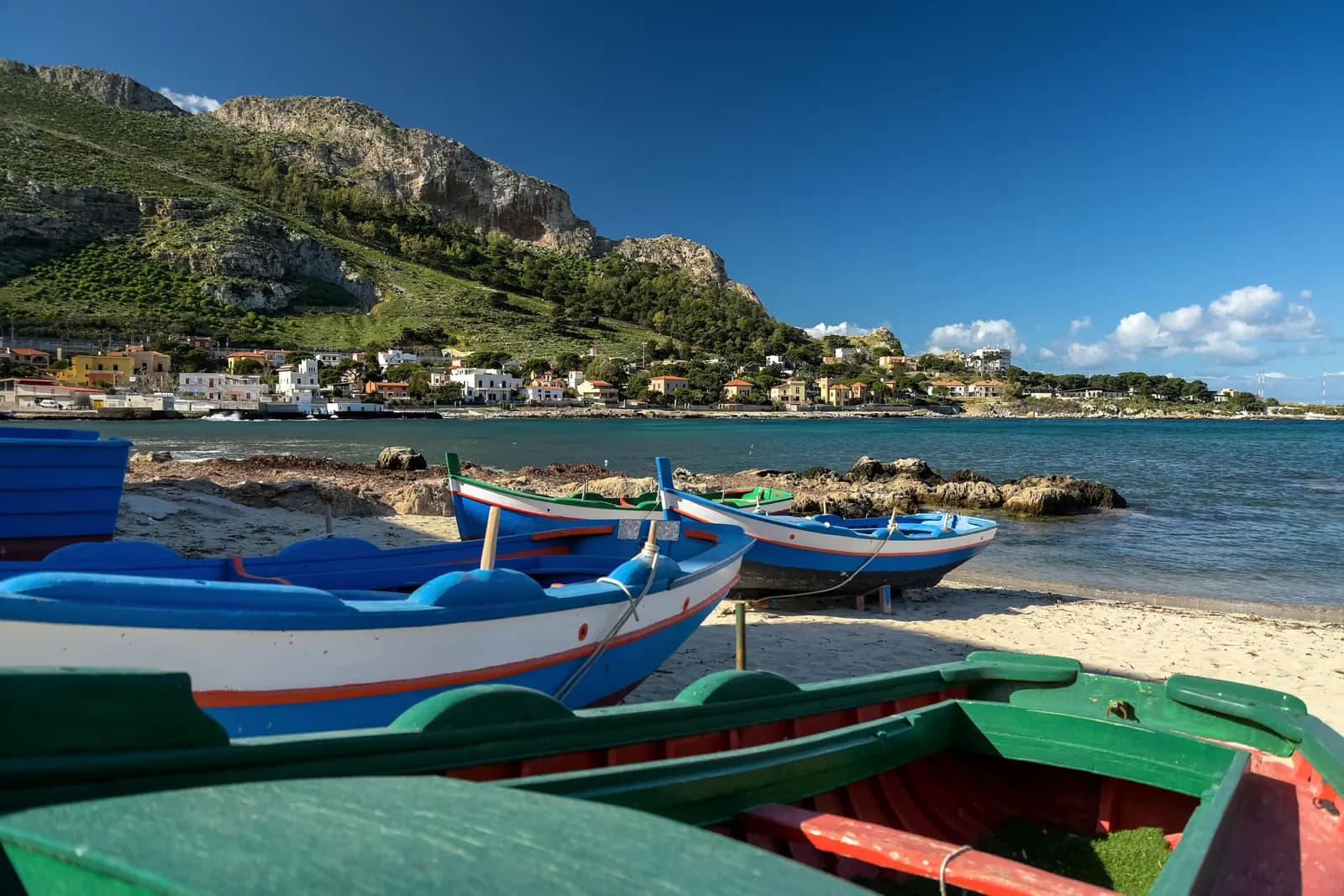 Painted wooden fishing boats pulled up on the sand at Sferracavallo, with the village and headland across the bay