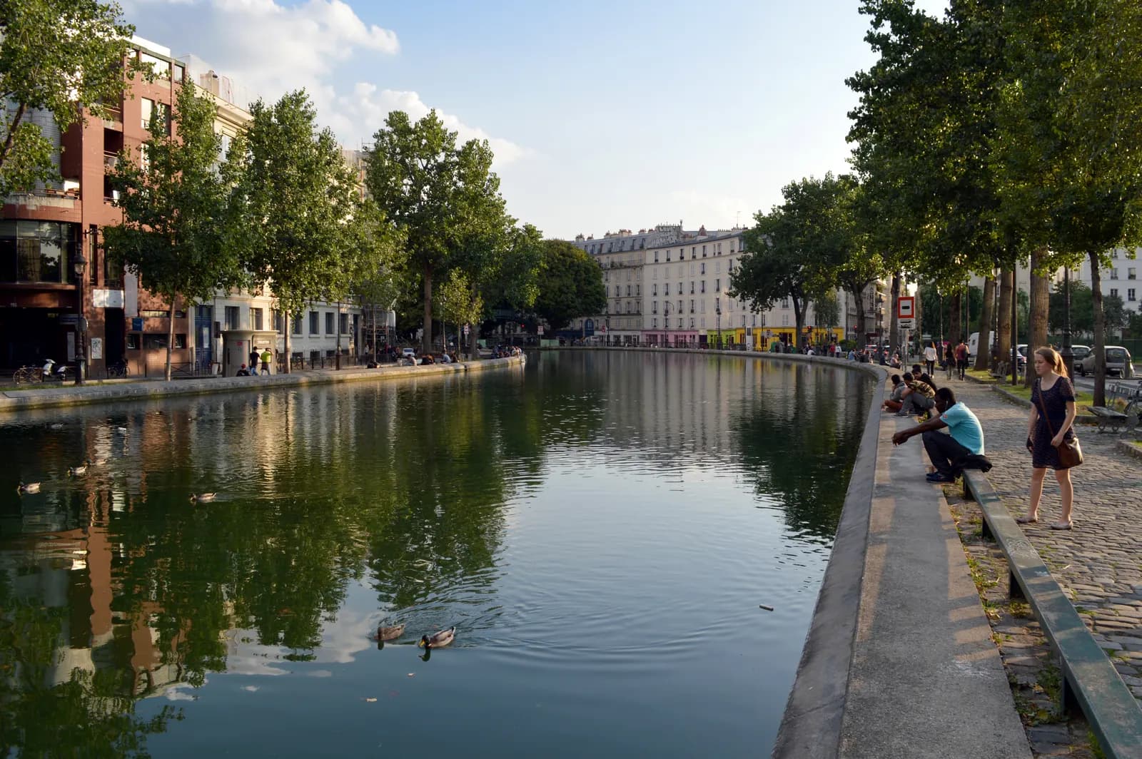 The Canal Saint-Martin seen from the quai de Valmy, with plane trees along both banks, ducks on the water and people sitting on the edge