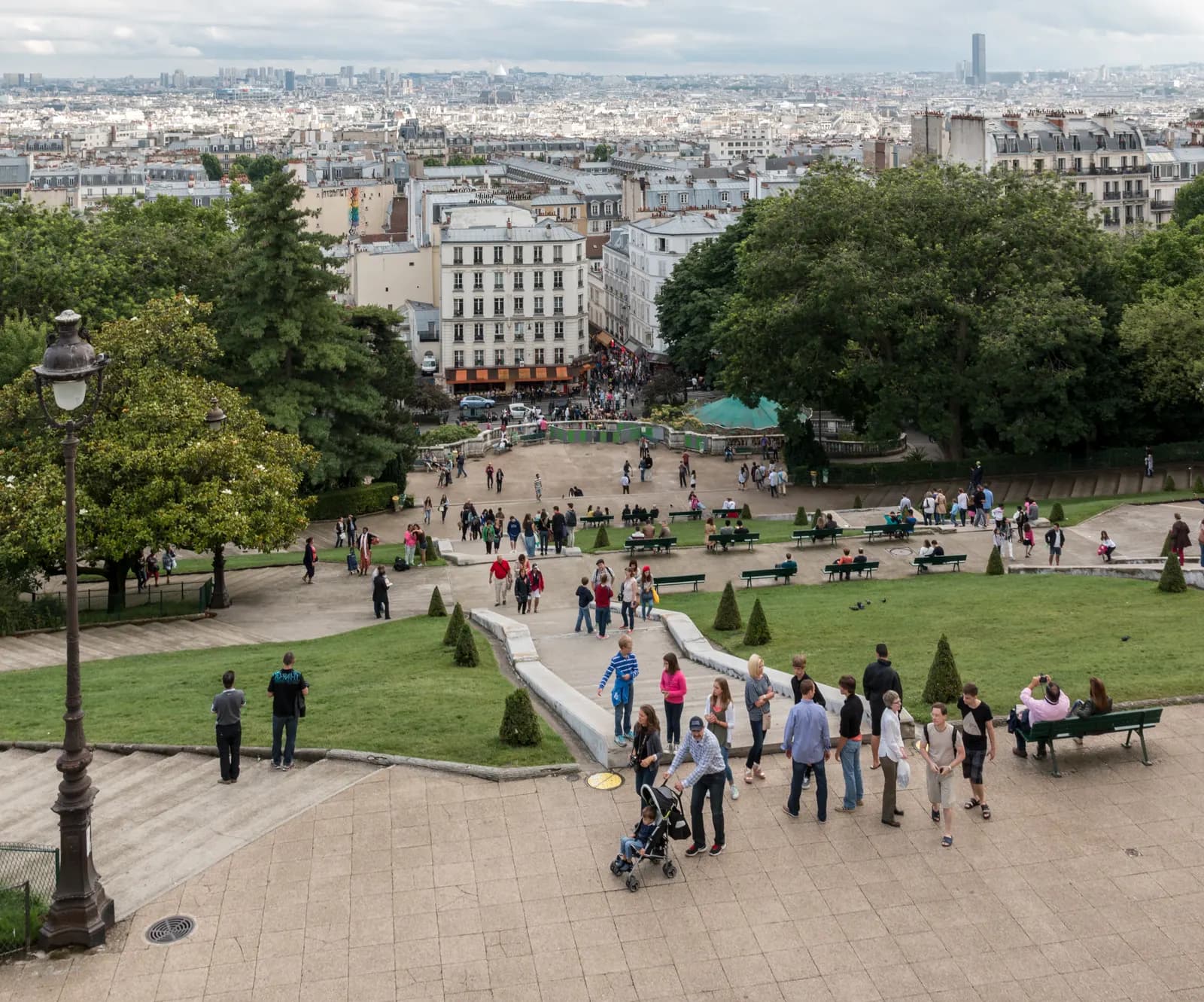 The rooftops of Paris seen from the terrace below the Sacré-Cœur basilica on the Montmartre hill, with visitors on the steps and the Montparnasse tower on the skyline