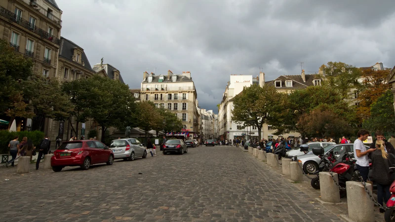 Cars parked along the cobbled place Saint-Germain-des-Prés at the foot of rue Bonaparte, with cafés and Haussmann buildings behind