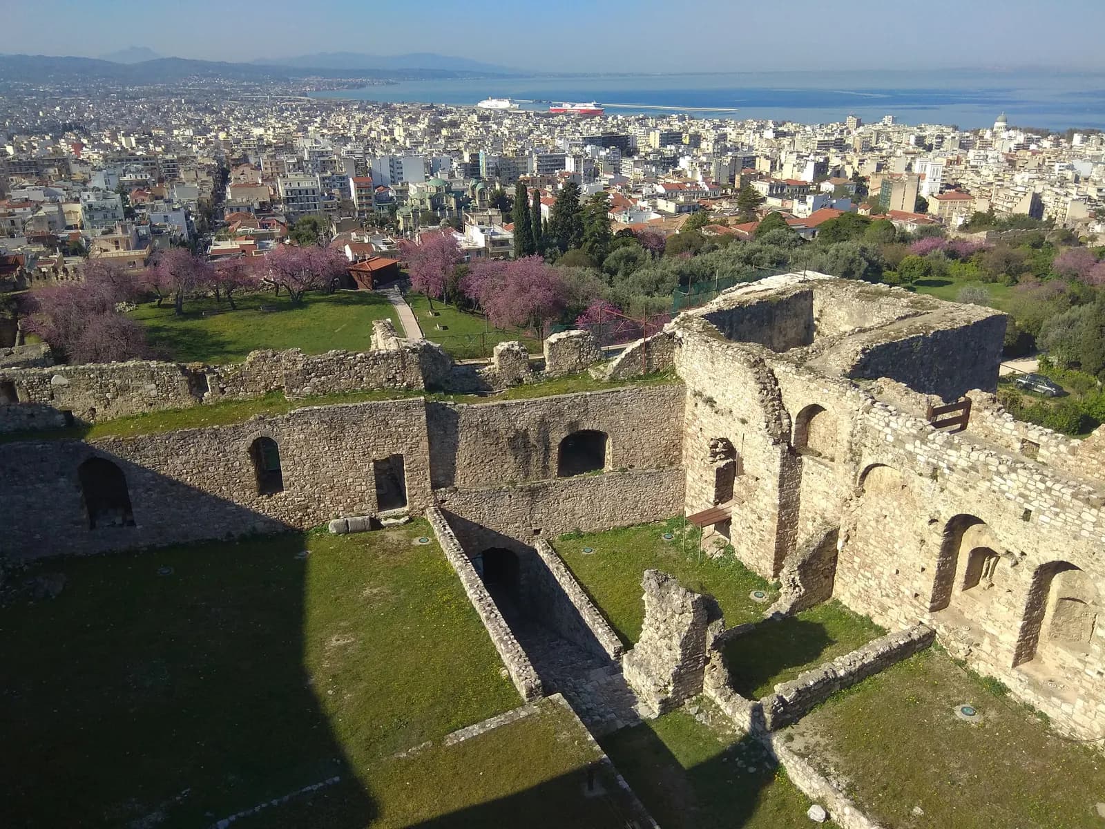 The stone walls and grassy courtyards of Patras Castle with trees in pink blossom, above the roofs of the city and the Gulf of Patras, with ships in the port