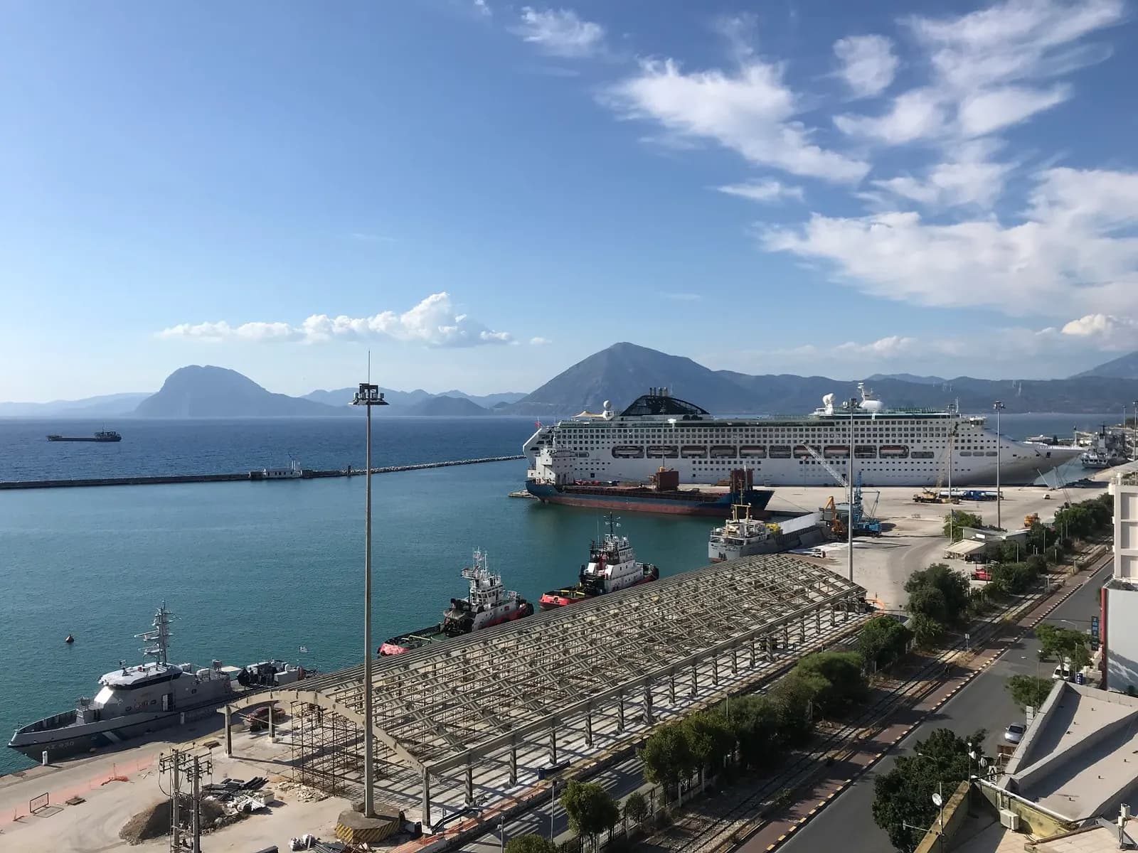 A cruise ship and tugboats moored in the port of Patras beside the seafront road, with the mountains across the gulf behind