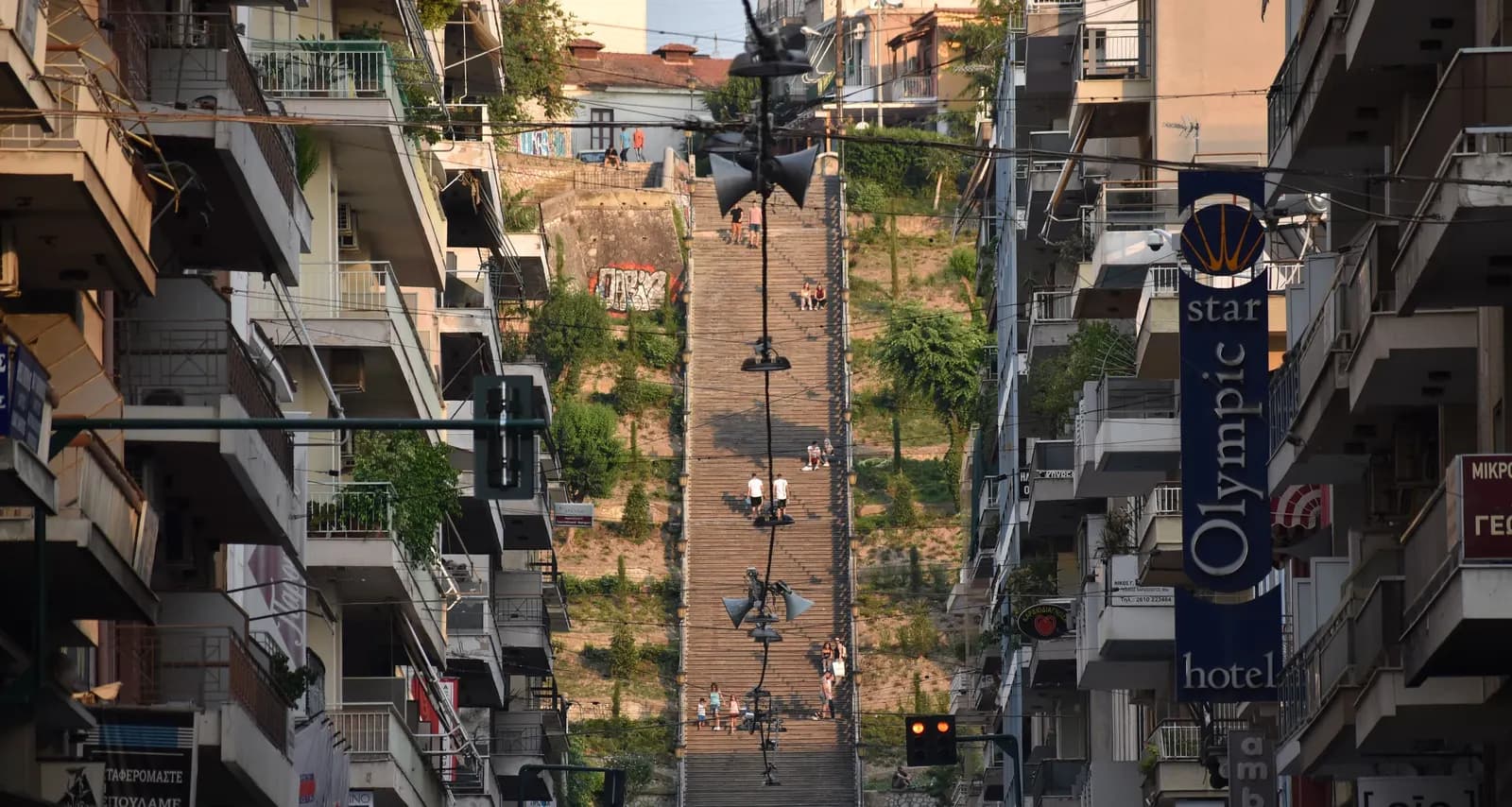 A long public stairway climbing a green hillside between apartment buildings and shop signs in central Patras, with a few people on the steps