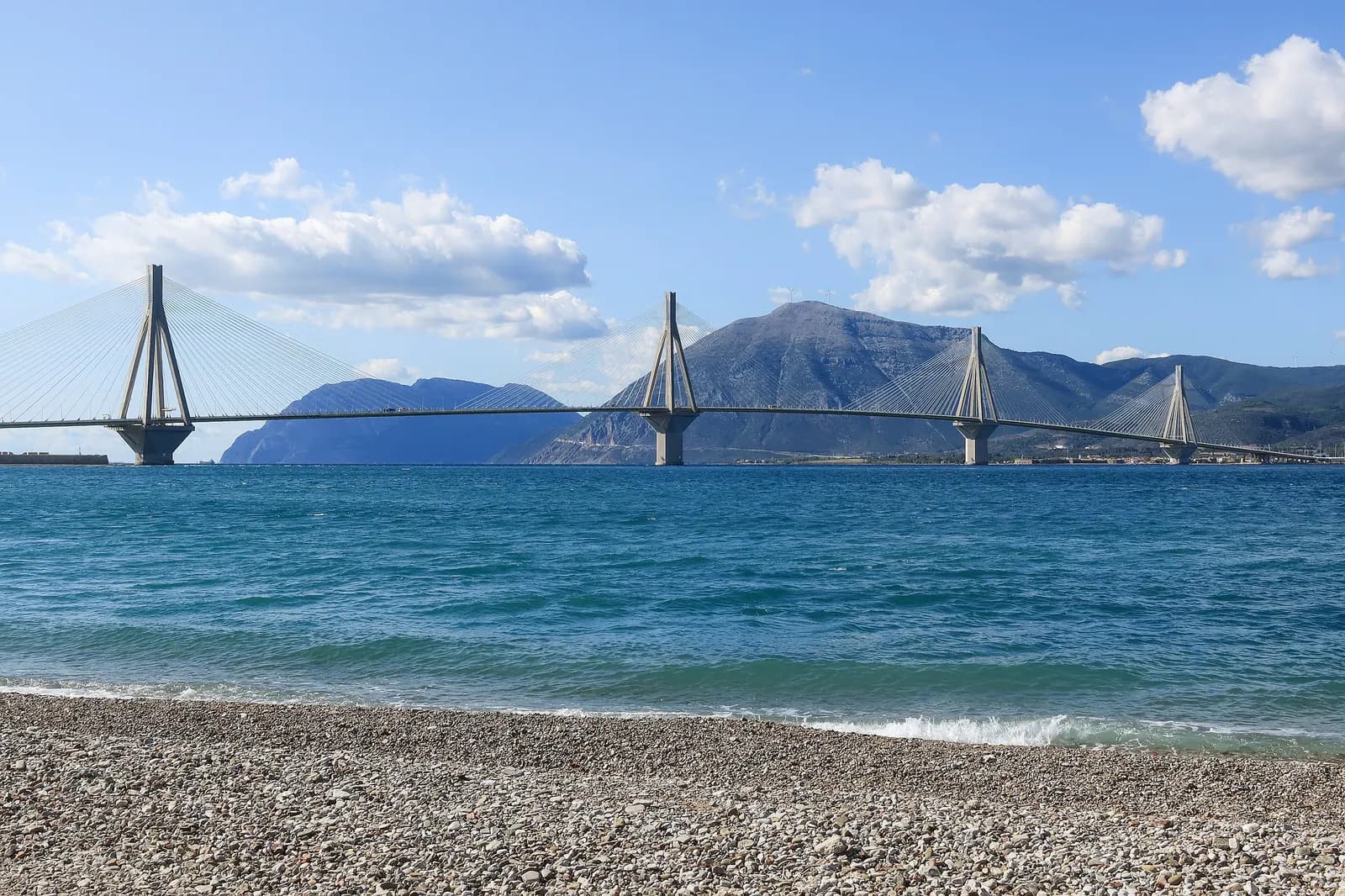 The Rio-Antirrio cable-stayed bridge crossing the strait from a pebble beach at Rio, with mountains on the far shore and blue sky