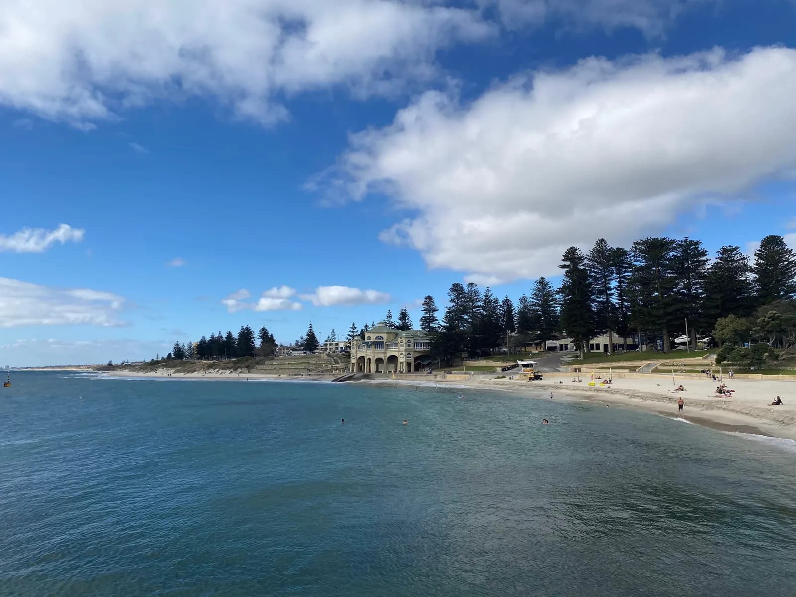 Cottesloe Beach seen from the water, with swimmers, people on the sand, a beachfront pavilion building with arches and tall pines behind the beach
