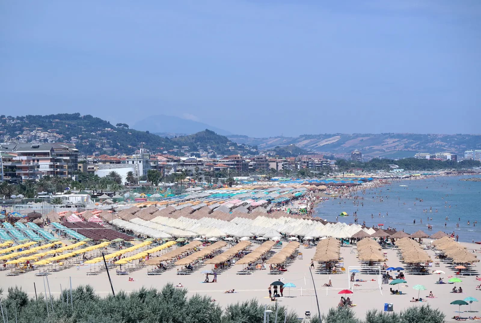 Pescara's sand beach seen along the shore, filled with rows of straw and coloured parasols and sunbeds, with apartment blocks behind and the Abruzzo hills in the distance.