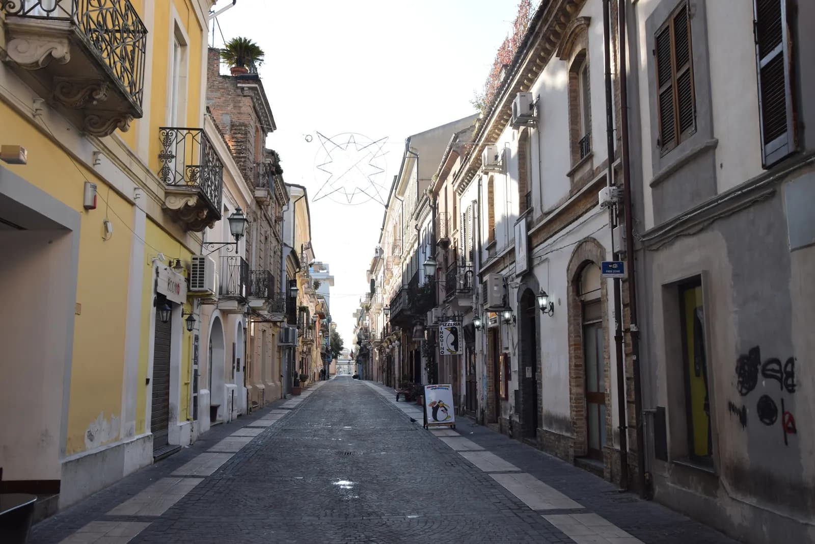 An empty paved street in Pescara Vecchia lined with low painted houses, wrought-iron balconies and shuttered bar fronts, with a star-shaped light strung across it.