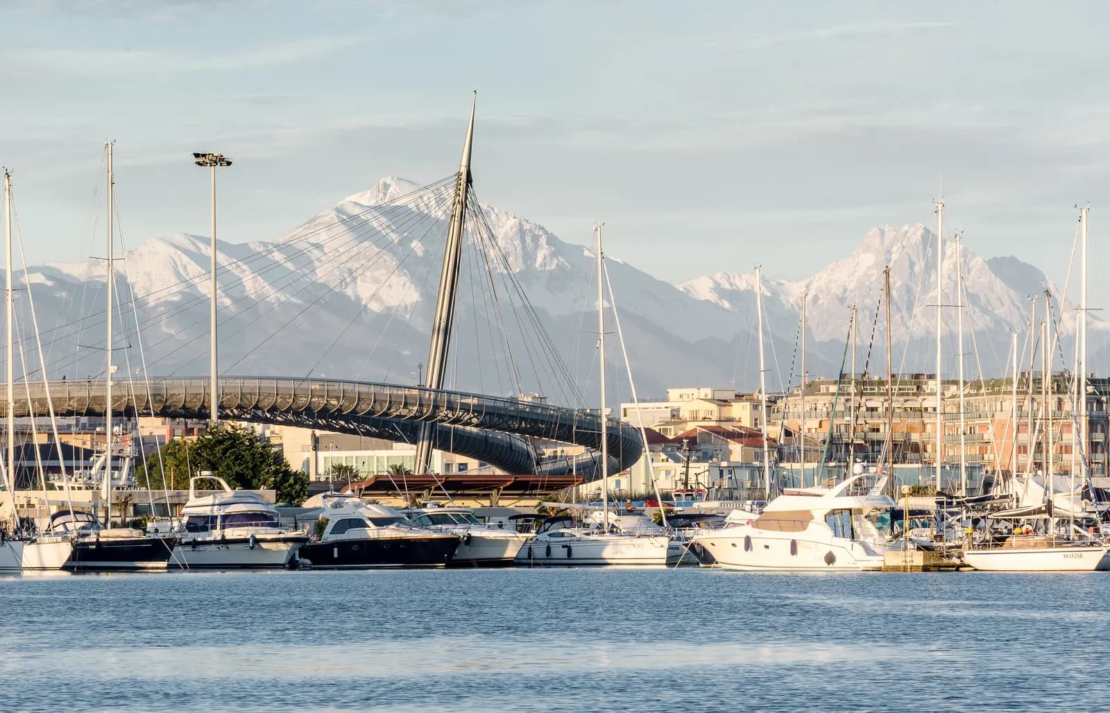 Yachts and motor boats moored in the Marina di Pescara, with the curved cable-stayed deck of the Ponte del Mare behind them and the snow-covered peaks of the Gran Sasso on the horizon.