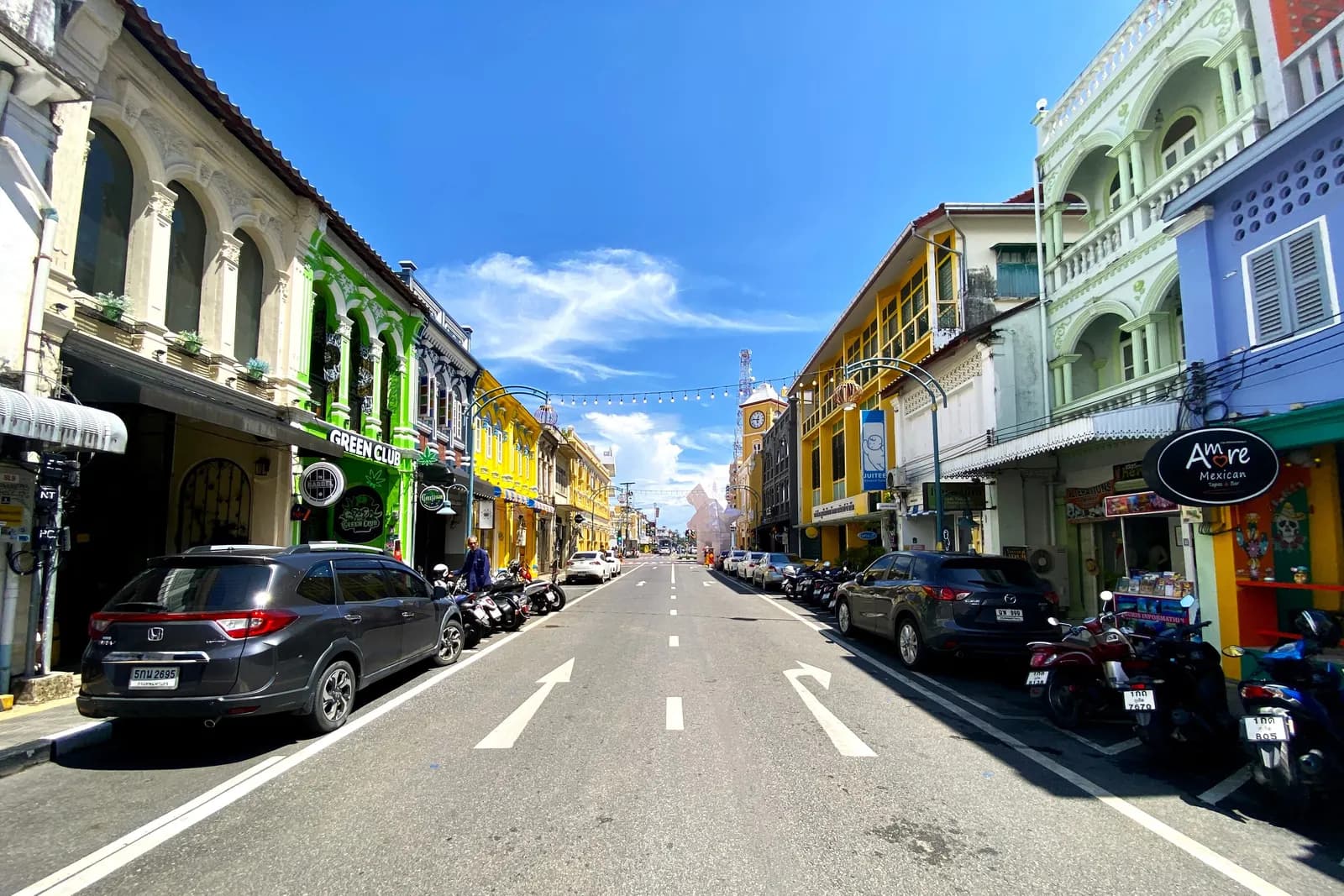 Phang Nga Road in Old Phuket Town, lined with colourful shophouses, with cars and motorcycles parked along both kerbs and a clock tower at the end of the street
