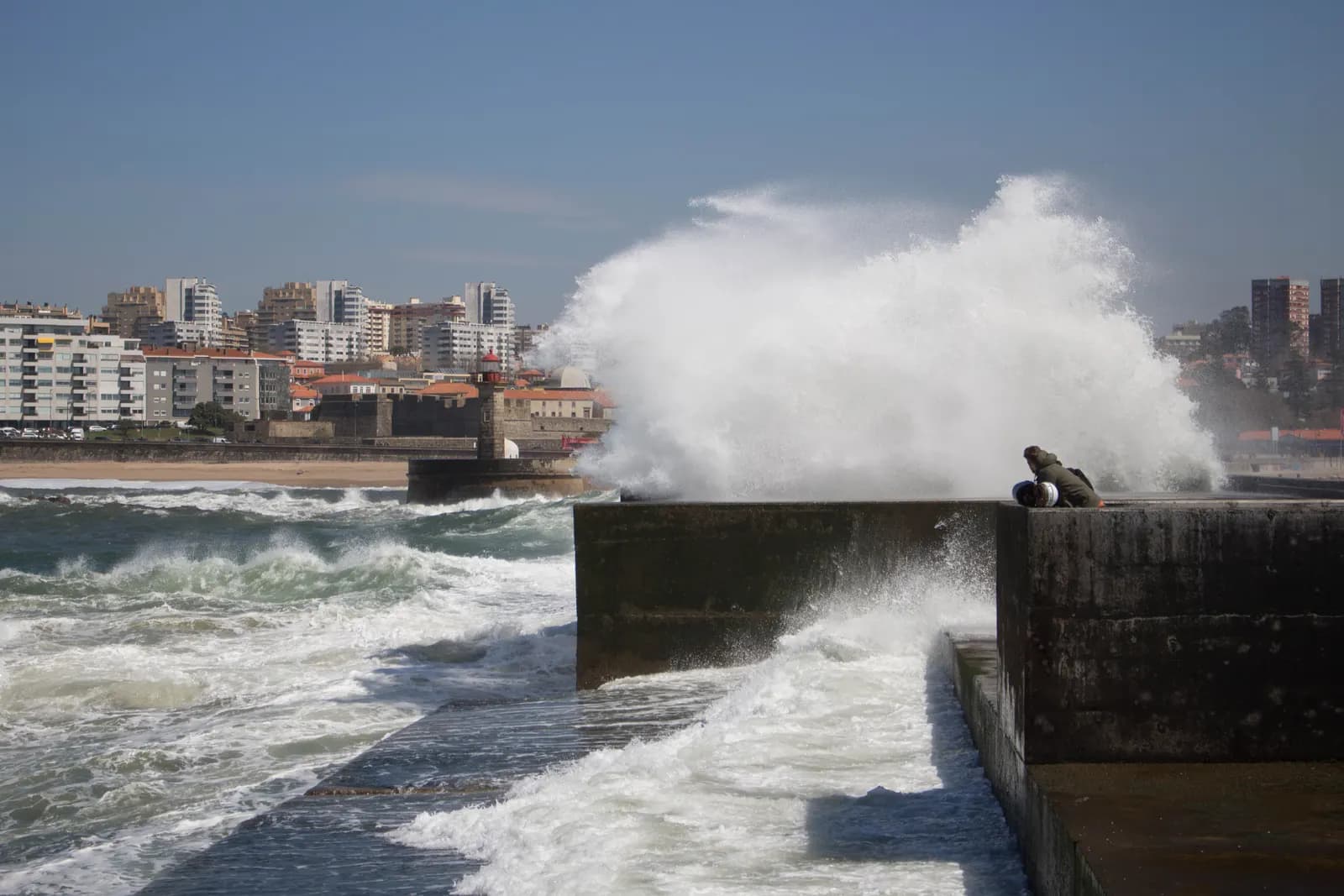 An Atlantic wave breaking over the pier at Foz do Douro, with apartment blocks along the seafront behind.