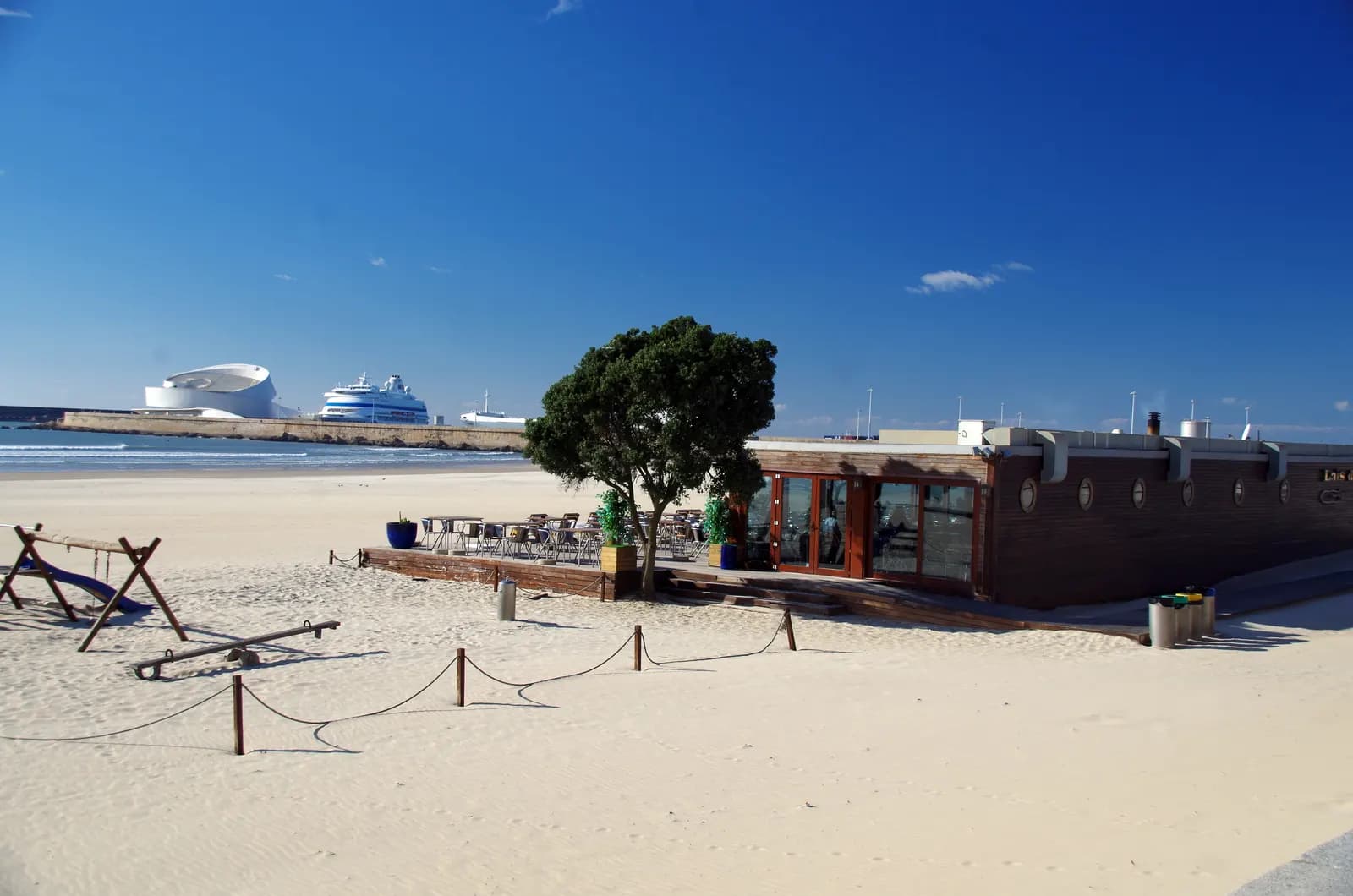 Wide sandy Matosinhos beach with a beach bar, a playground and the Leixões cruise terminal in the distance.
