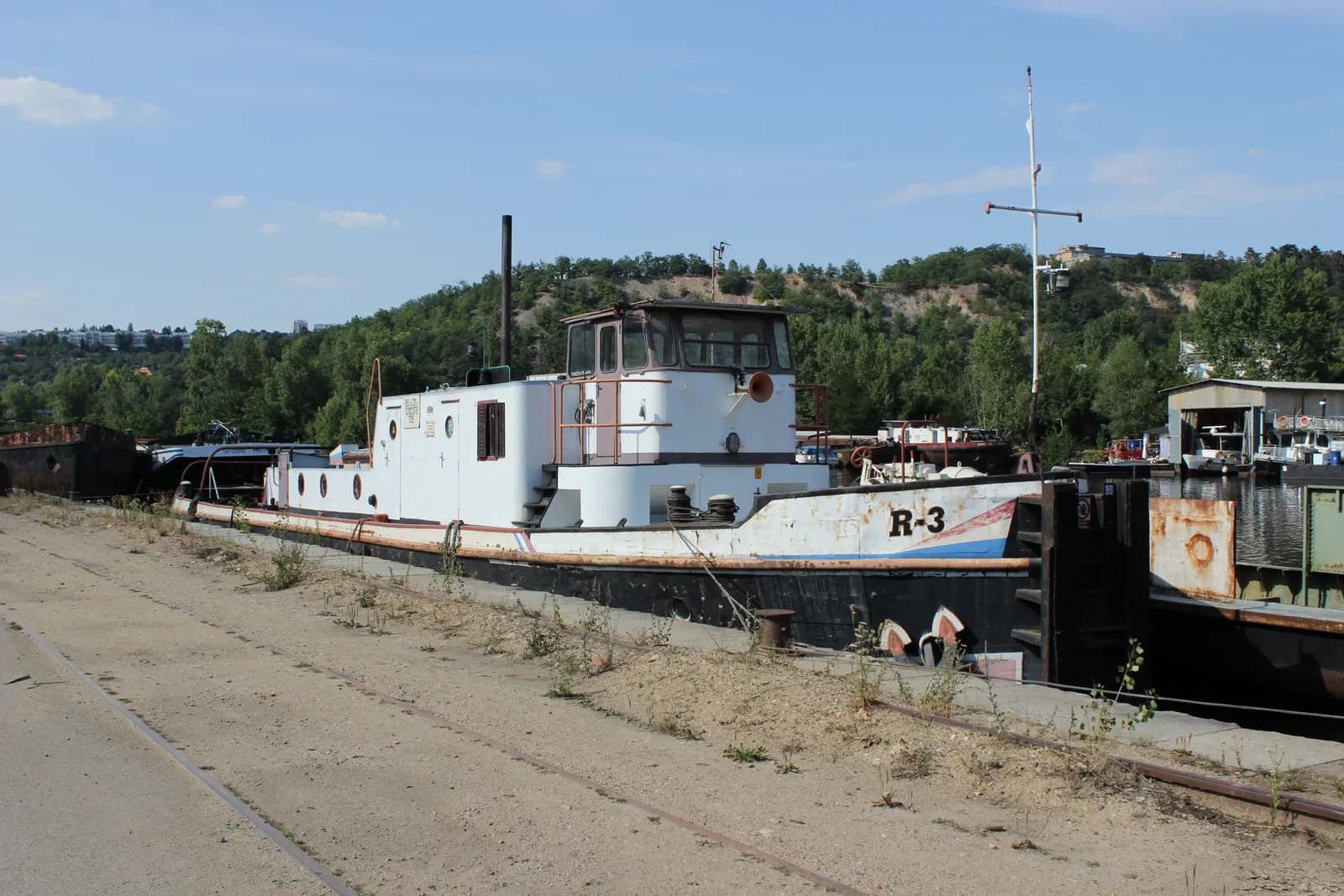 An old white river boat marked R-3 moored at the Holešovice docks in Prague, beside an old quay with rails and a wooded hill behind