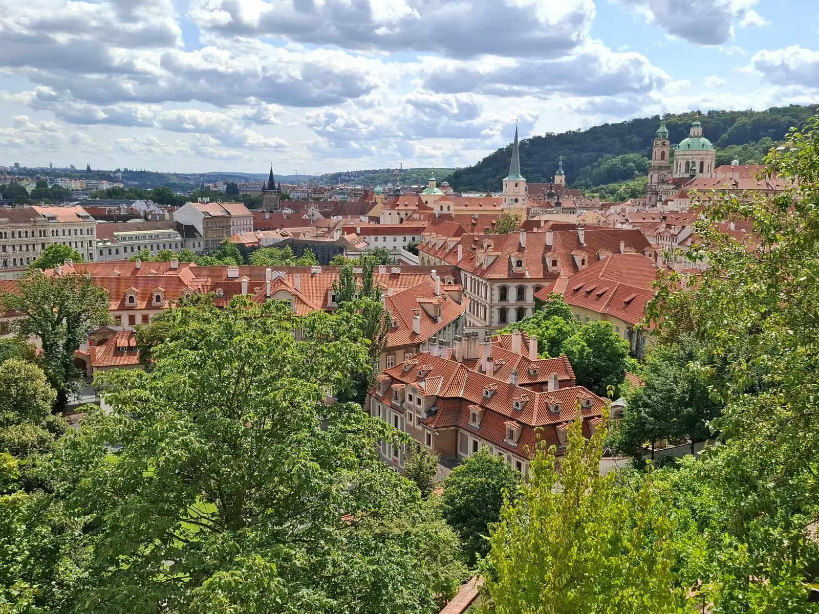 Red tiled roofs of Malá Strana seen from above between green trees, with church towers and a wooded hill behind
