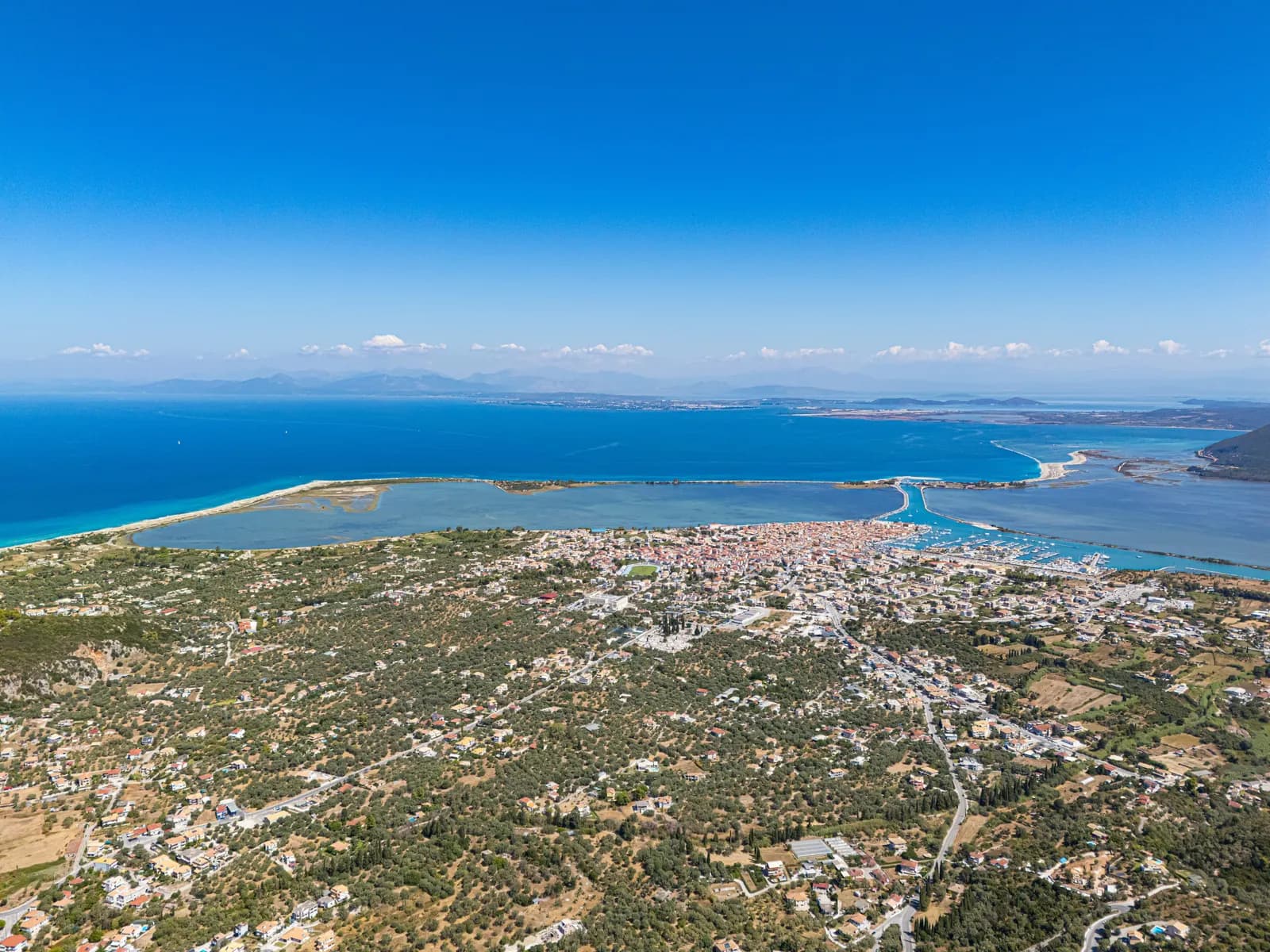 Aerial view of Lefkada Town among olive groves, with the lagoon, the causeway and the marina beyond it and the mainland coast across the water