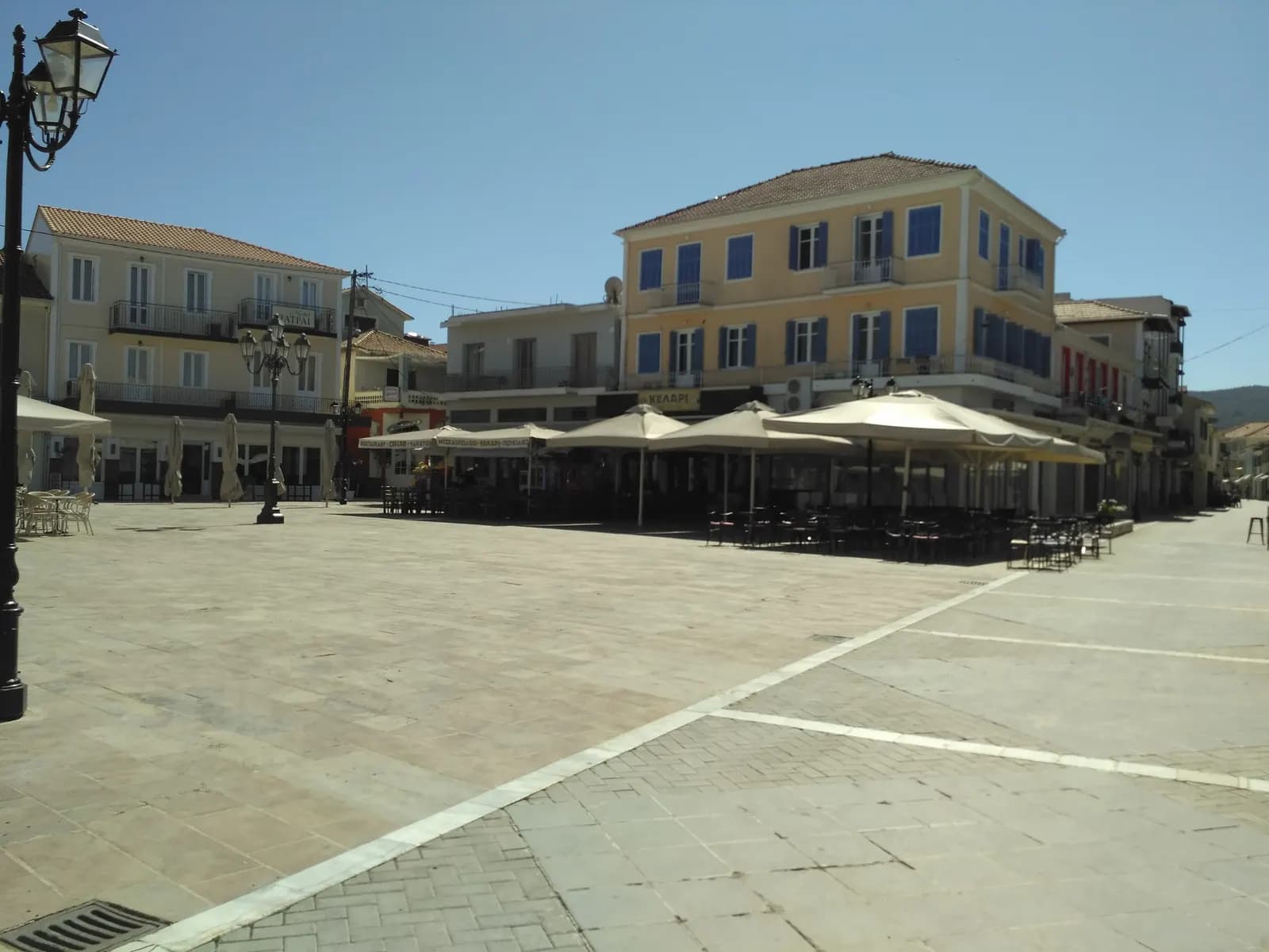 The paved main square of Lefkada Town with cafe parasols in front of pastel buildings with blue shutters, under a clear sky