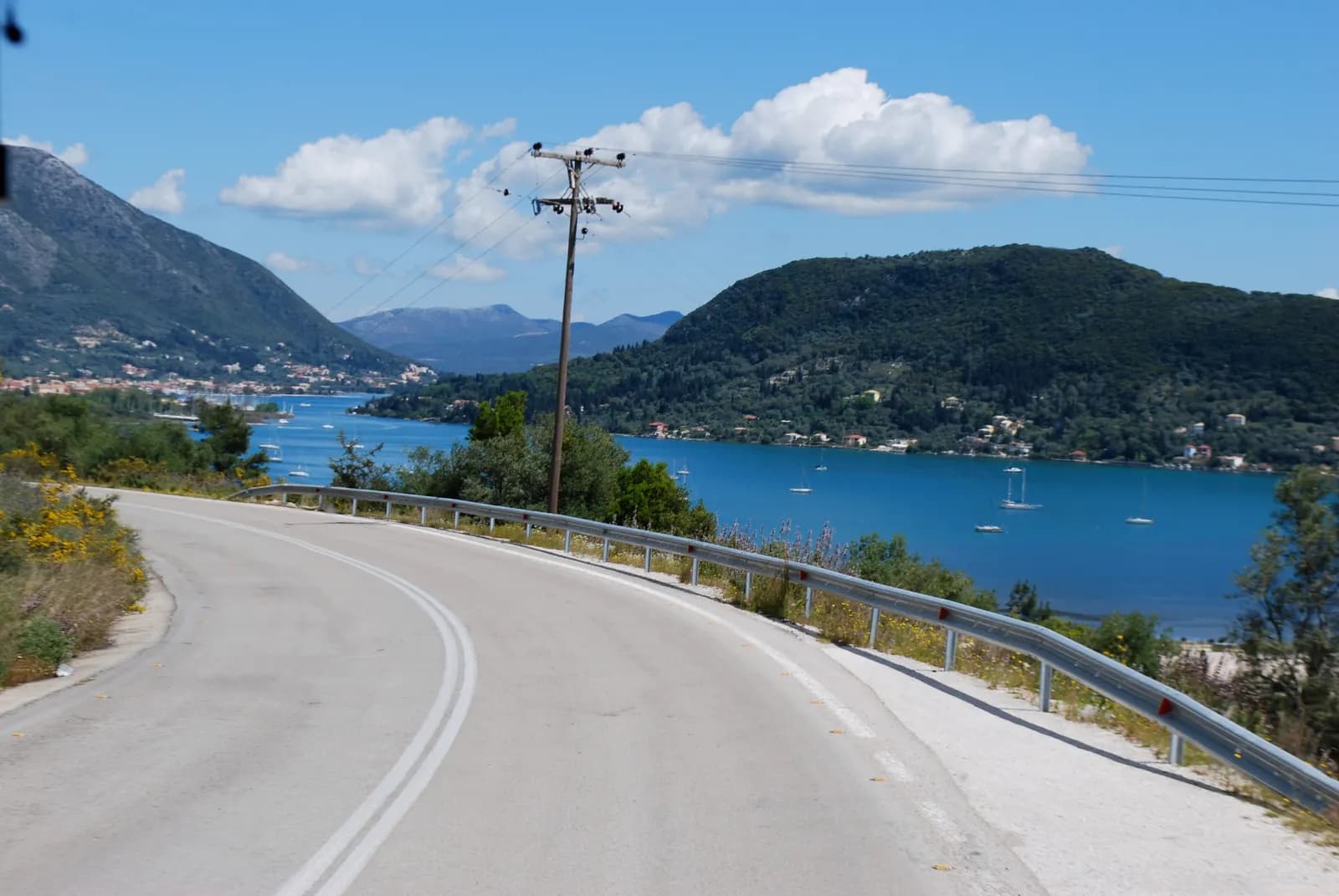 A road curving above the bay of Nydri on the east coast of Lefkada, with sailing boats at anchor, the town on the shore and wooded hills across the water