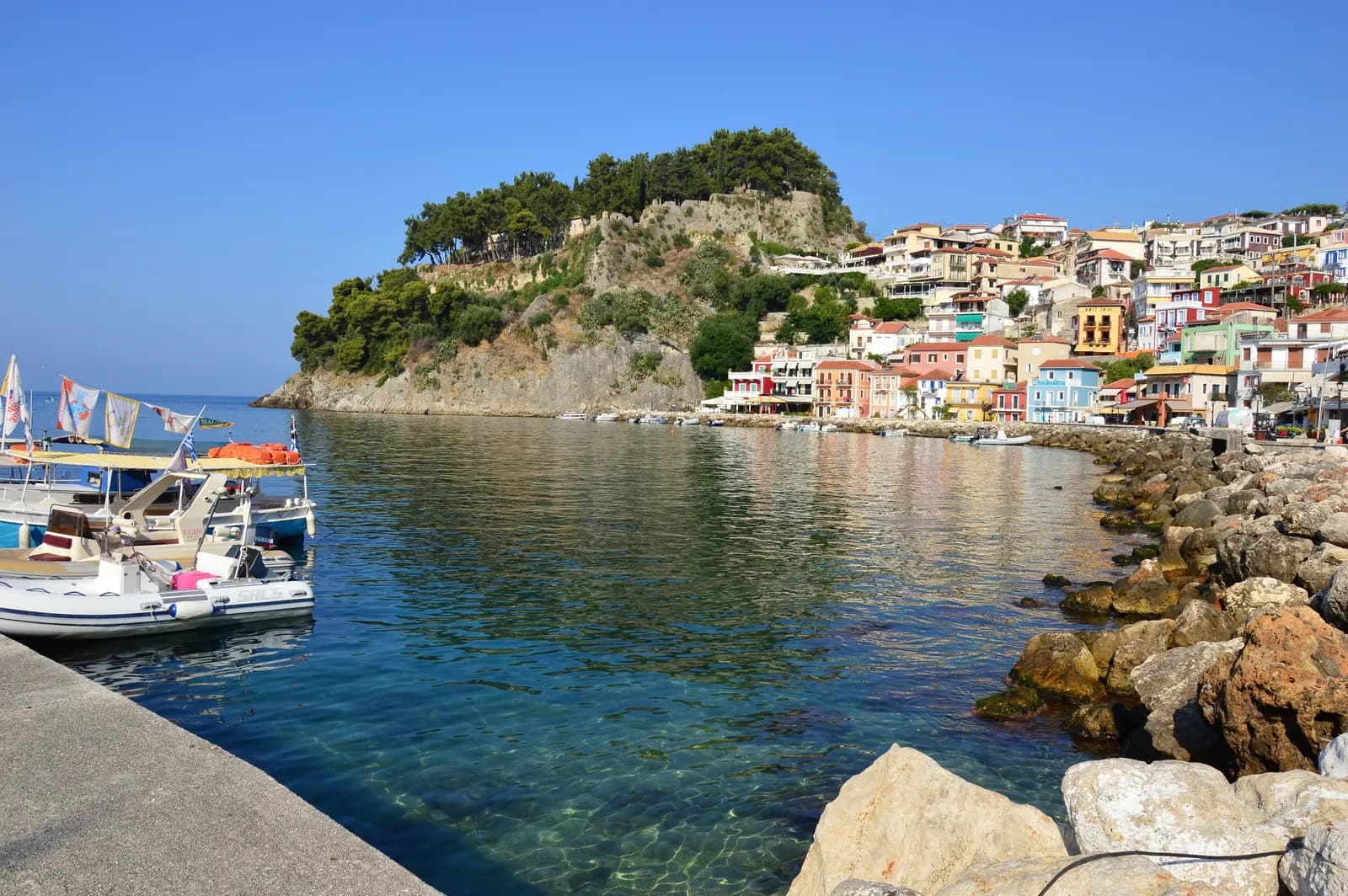 The bay of Parga with clear water by the harbour wall and rocks, the castle hill covered in pines and colourful houses climbing the slope
