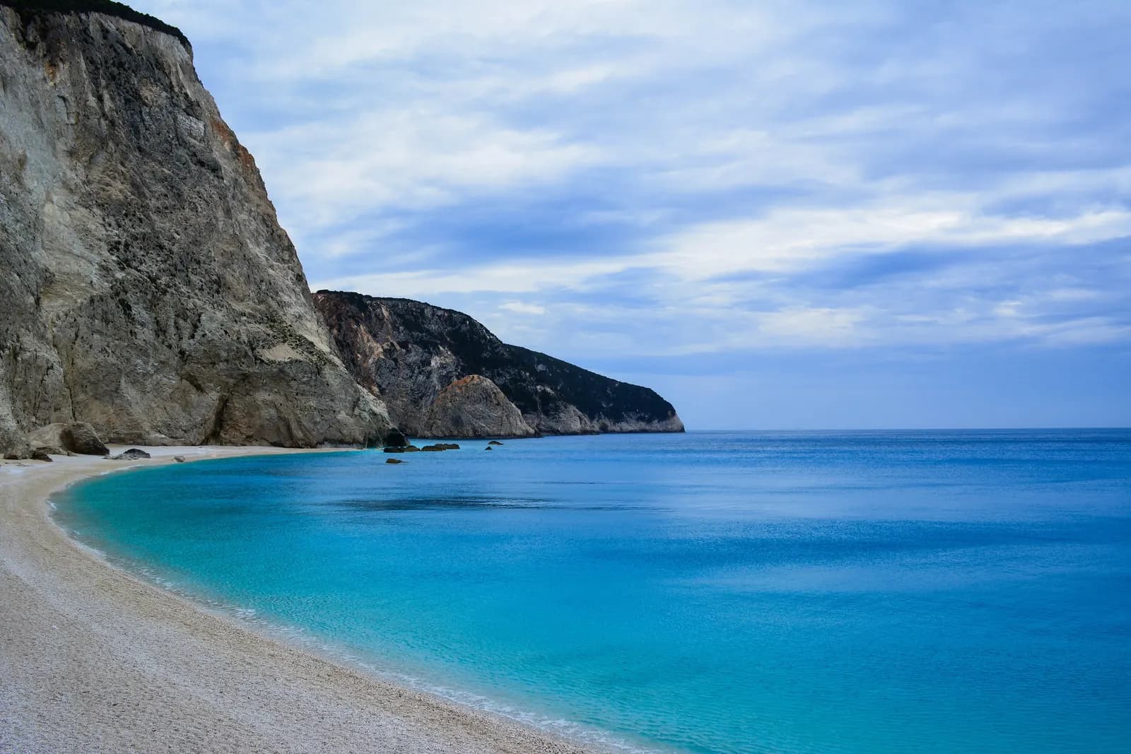 The pale pebble beach of Porto Katsiki on the west coast of Lefkada, curving below tall white limestone cliffs, with turquoise water and a cloudy sky