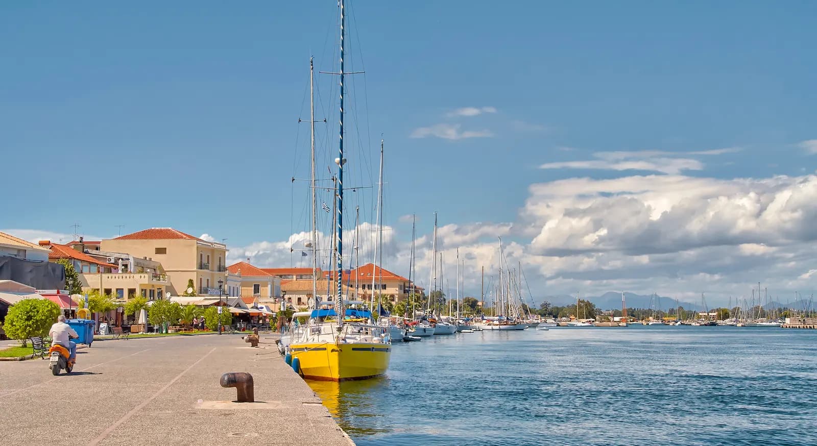 The waterfront promenade of Preveza with cafes under the trees, a yellow sailing boat and a line of yachts moored along the quay on a sunny day