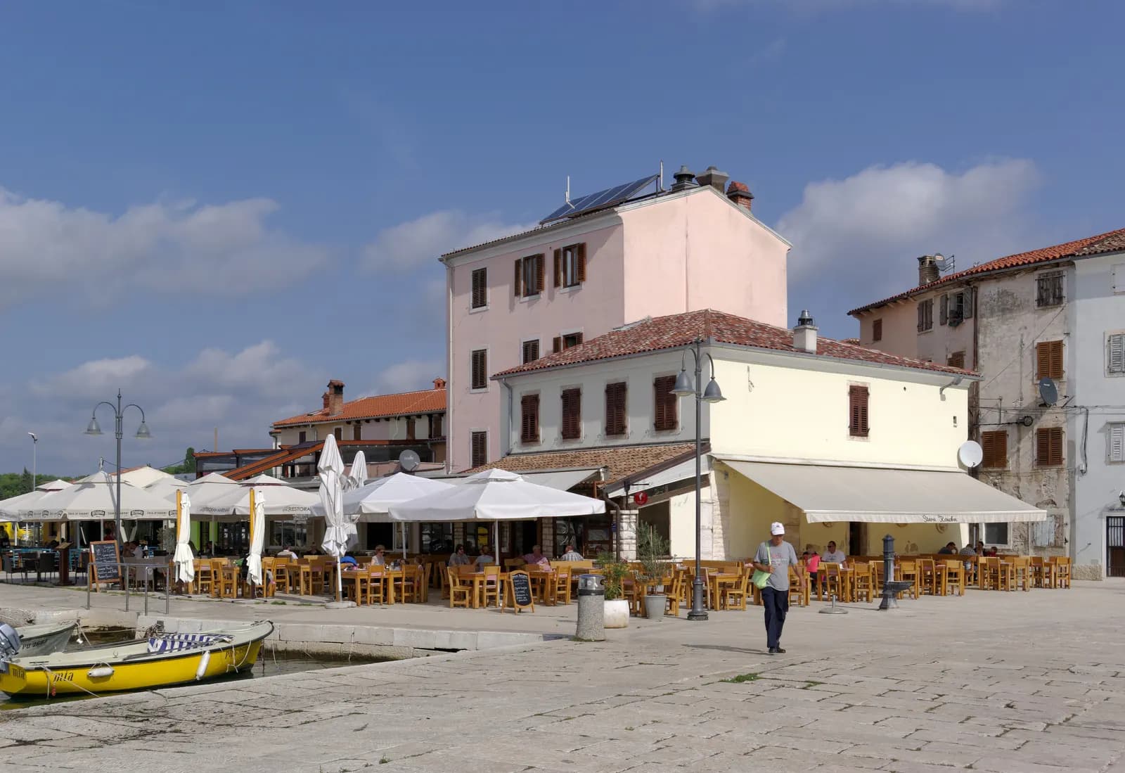 Restaurant terraces along the stone quay of Fažana harbour, with a small yellow boat moored in the foreground