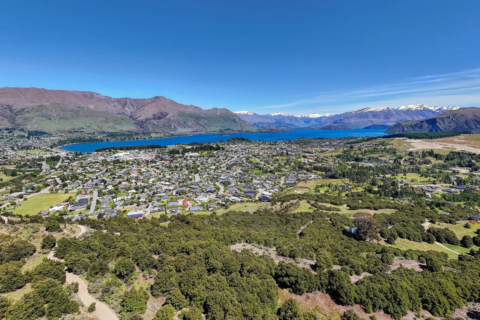 Houses of Wānaka spreading towards the blue water of Lake Wānaka, with brown hills and snow-capped mountains behind, seen from the air