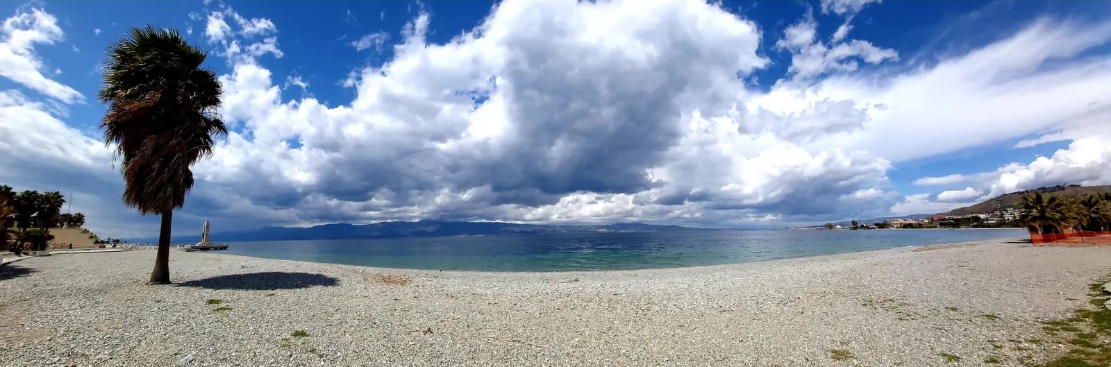 A wide pebble beach on the Reggio Calabria waterfront with a single palm tree, the Strait of Messina beyond it and the Sicilian coast under tall cloud.