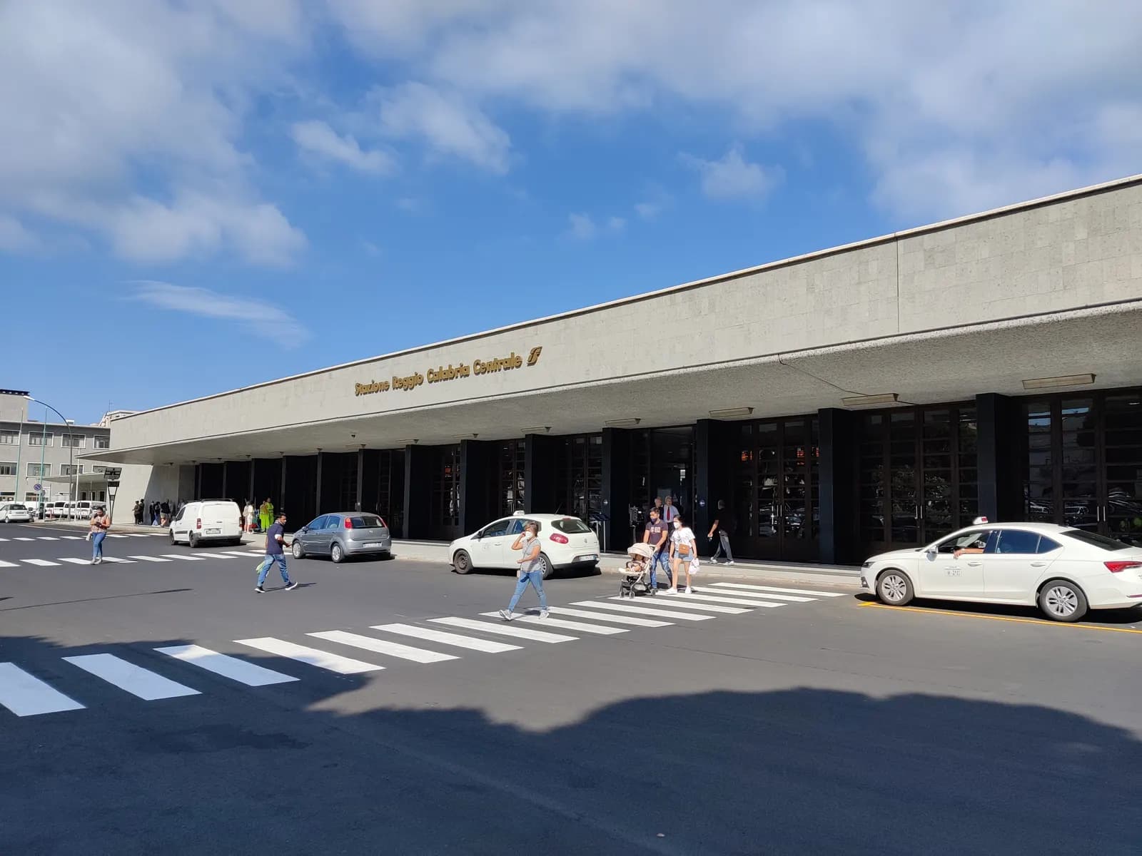 The long low concrete front of Reggio Calabria Centrale station, with its name in gold lettering, a zebra crossing, parked cars and a white taxi outside.