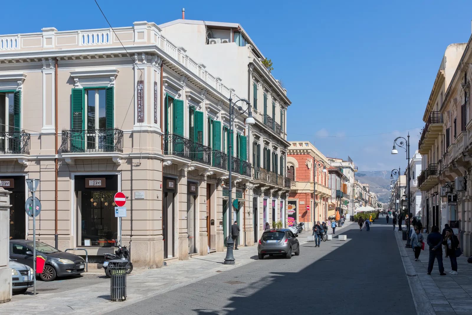Corso Giuseppe Garibaldi in Reggio Calabria, a straight shopping street of cream buildings with green shutters and iron balconies, with a few parked cars and people walking.