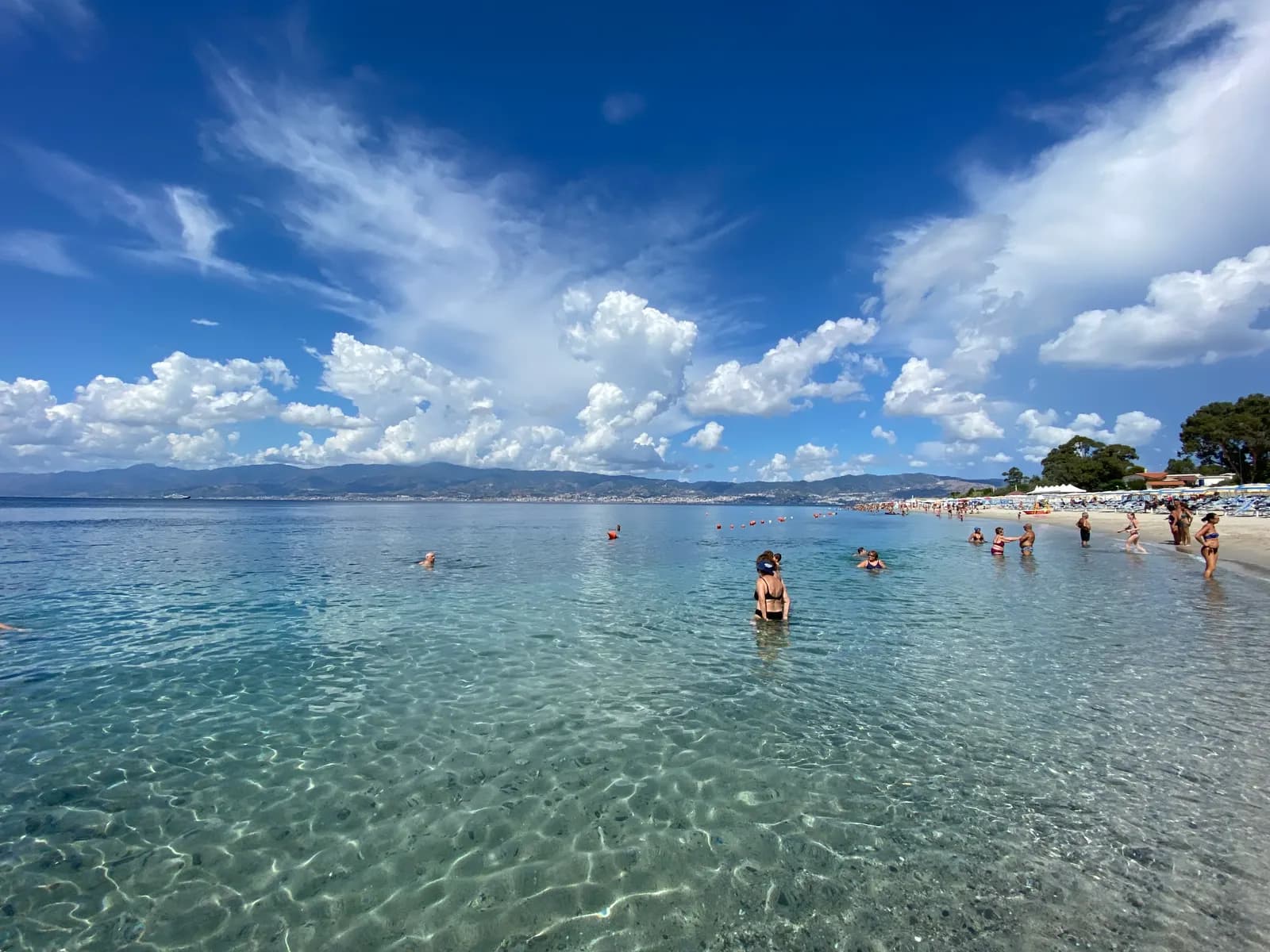 Shallow clear water off the beach at Catona north of Reggio Calabria, with bathers, parasols along the shore and the Sicilian coast across the Strait.