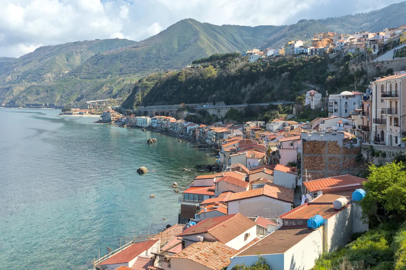 The fishing quarter of Chianalea at Scilla, its tiled roofs and tall houses packed along the water below a green headland, with the coast road and railway running behind.