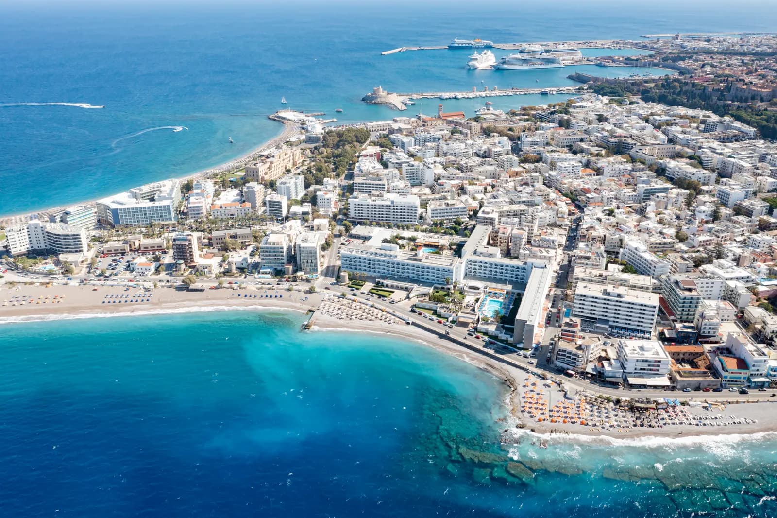 Aerial view of the Rhodes New Town seafront, with hotels behind a sand beach, the grid of town streets and the harbour with cruise ships beyond