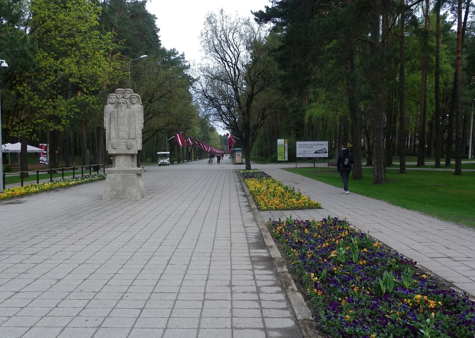 A paved alley through the pines of Mežaparks in Riga, with flower beds, a stone sculpture and Latvian flags