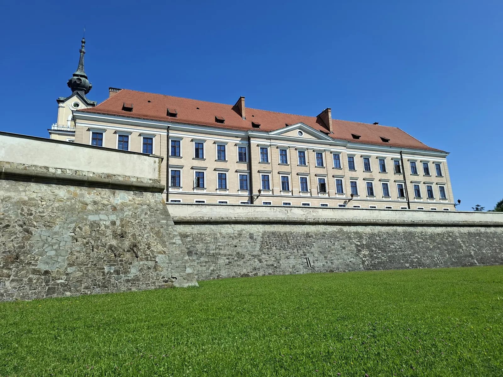 The Lubomirski Castle in Rzeszów, a long building with a red roof and a tower above stone fortification walls and a lawn